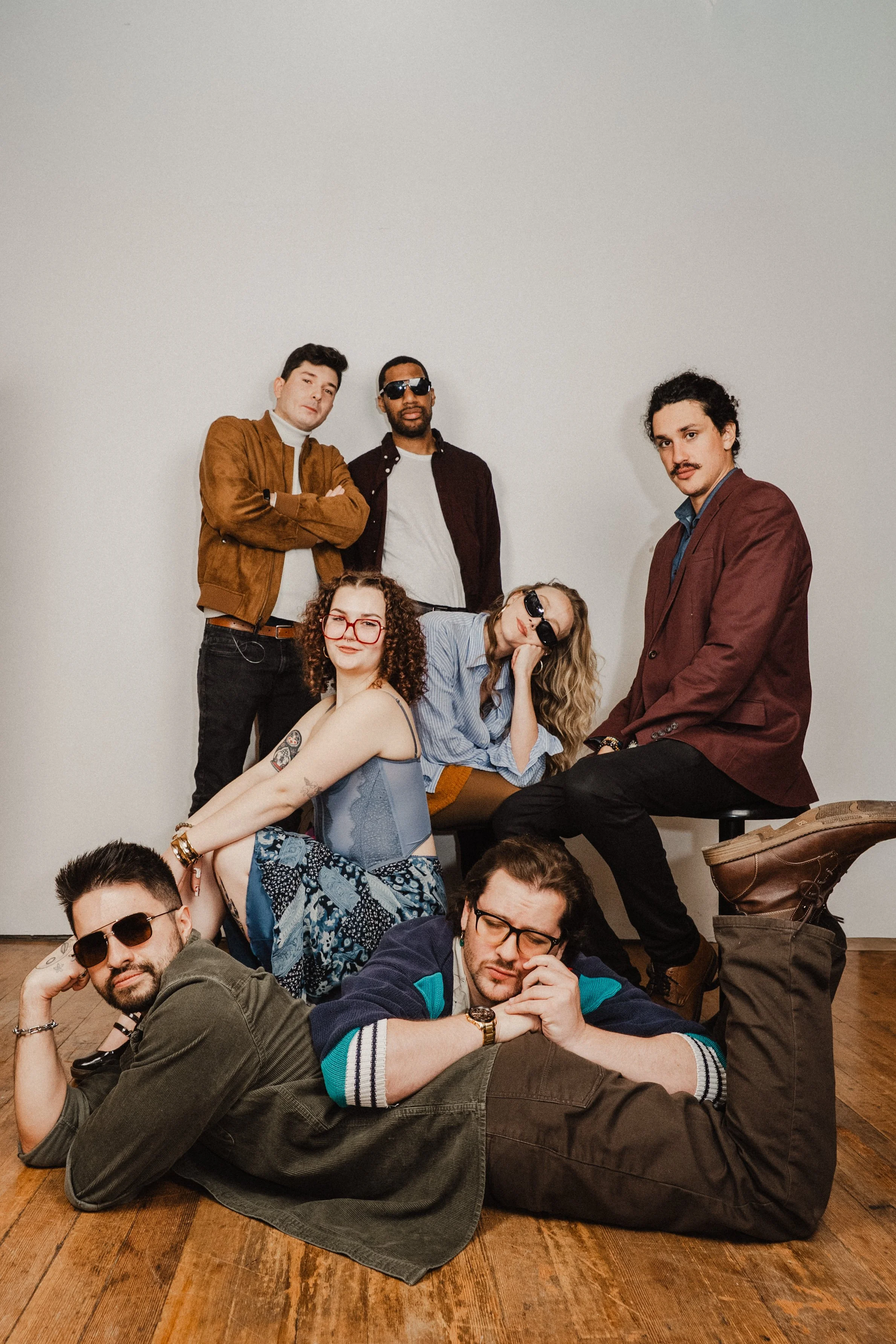 Group of seven young adults posing together indoors against a plain wall, some sitting and some standing, with a variety of casual and stylish clothing and accessories.