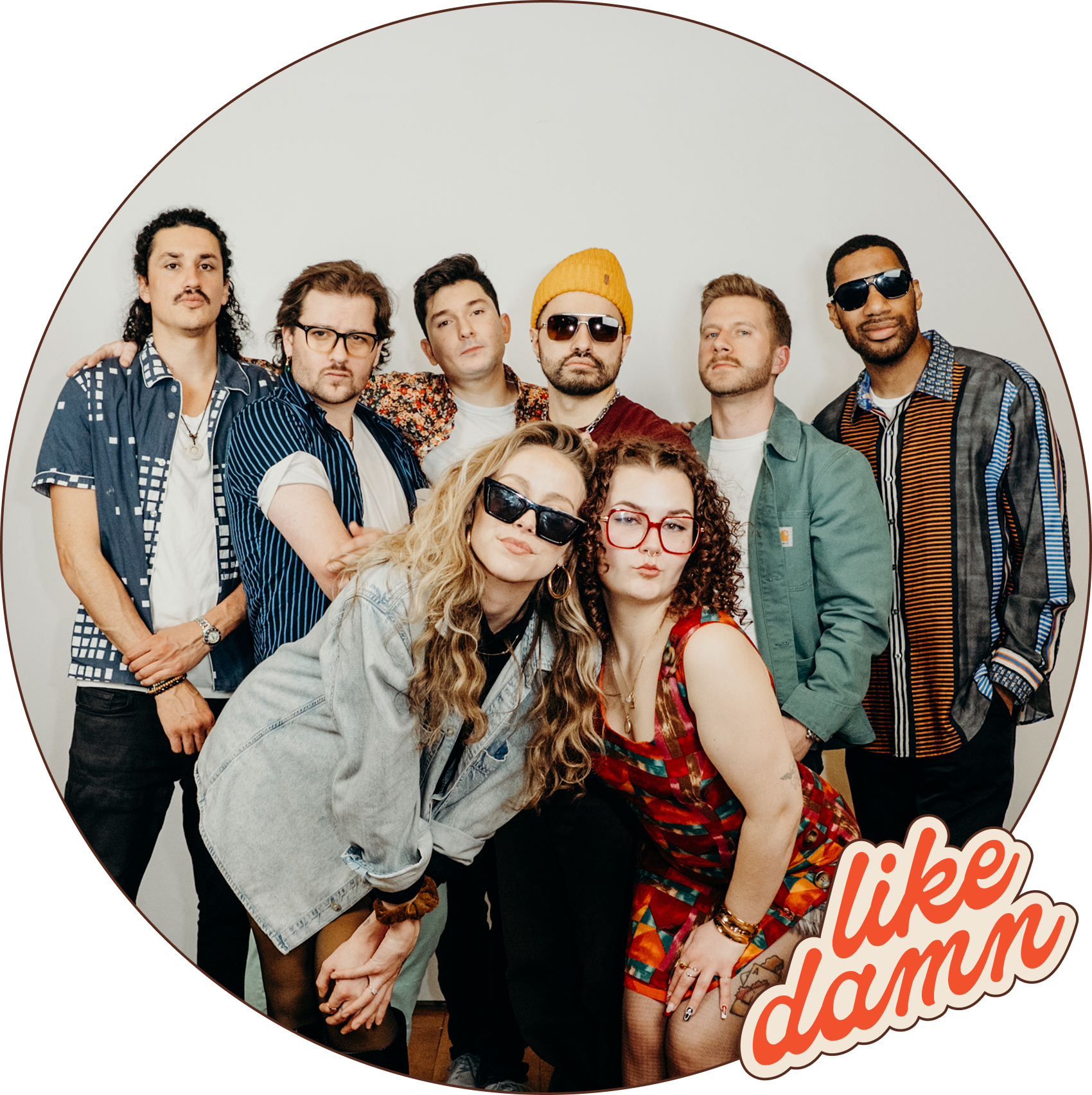 A group of nine diverse young adults posing together against a plain white background, some wearing sunglasses and colorful clothing, with the words 'like damn' written in stylized text at the bottom right corner.