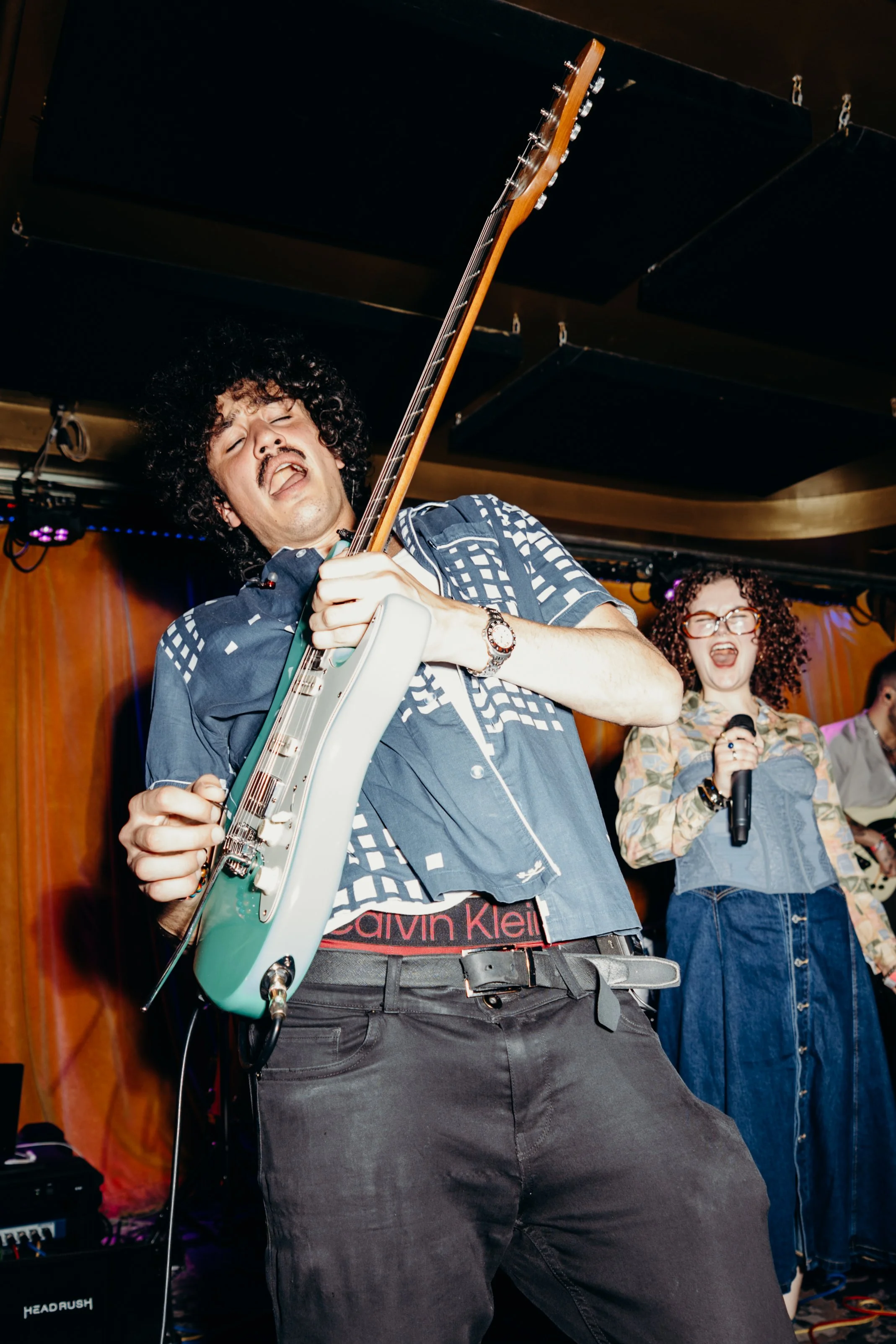 A man with curly hair and a mustache passionately plays an electric guitar during a live performance, with a woman singing into a microphone behind him.
