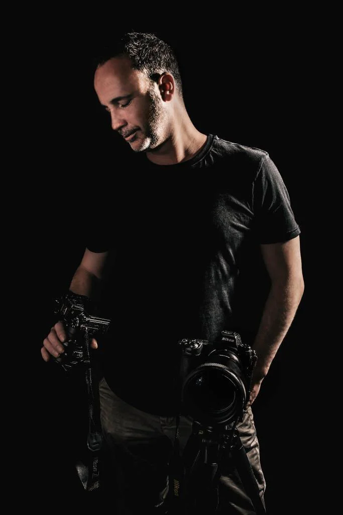 Photograph of a man in a black t-shirt holding two cameras against a dark background.