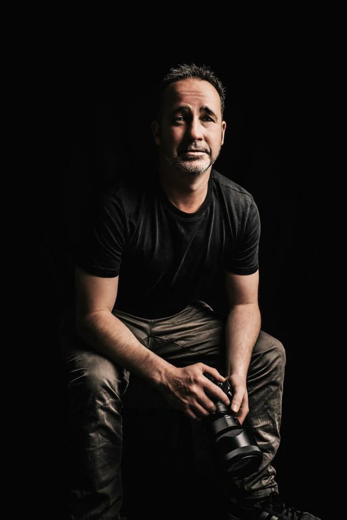 A man sitting against a black background, holding a camera in one hand, wearing a black t-shirt and camouflage pants, with a neutral expression.