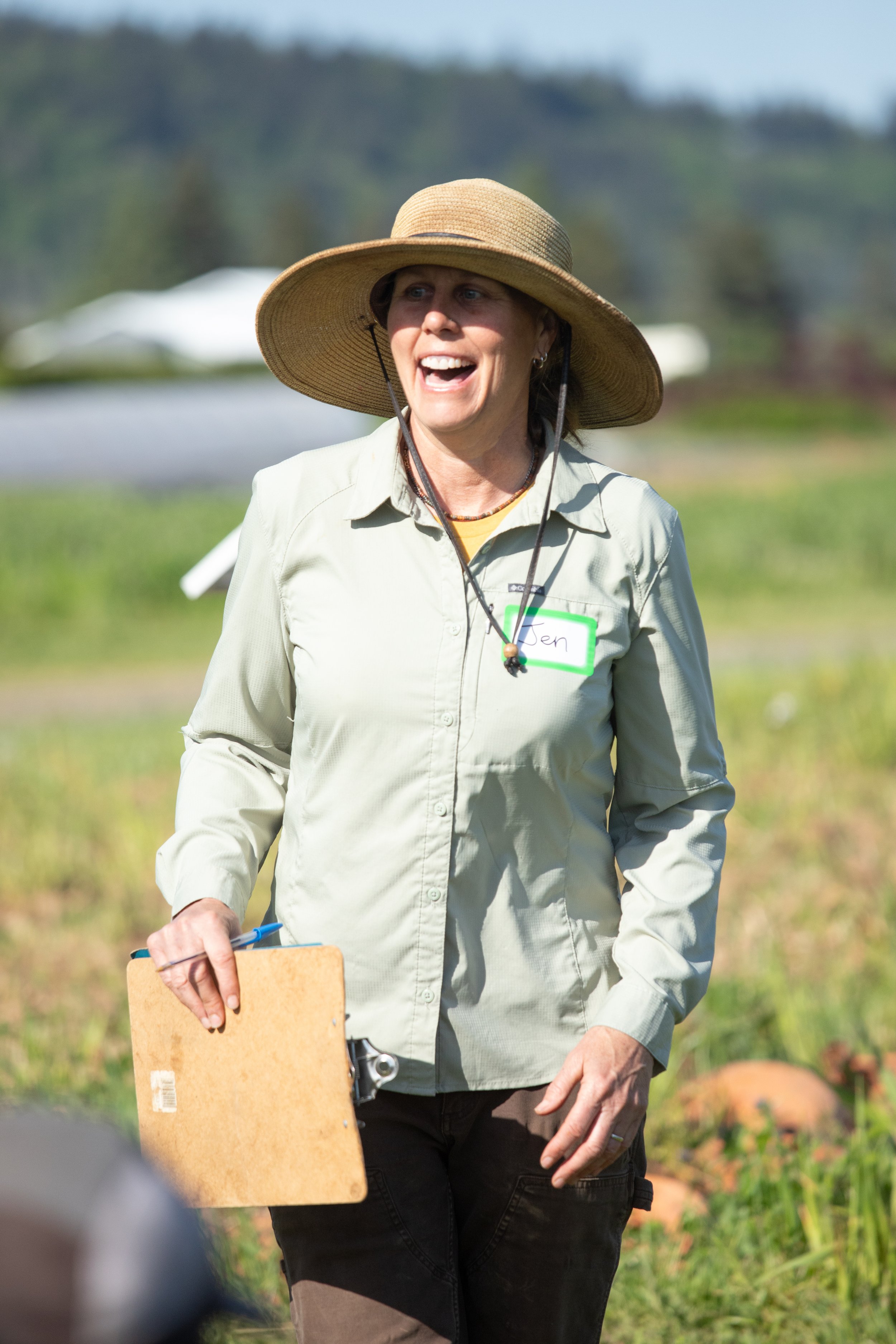 A woman wearing a wide-brimmed straw hat, a light green button-up shirt, and dark pants, smiling and holding a clipboard and pen outdoors in a natural setting.