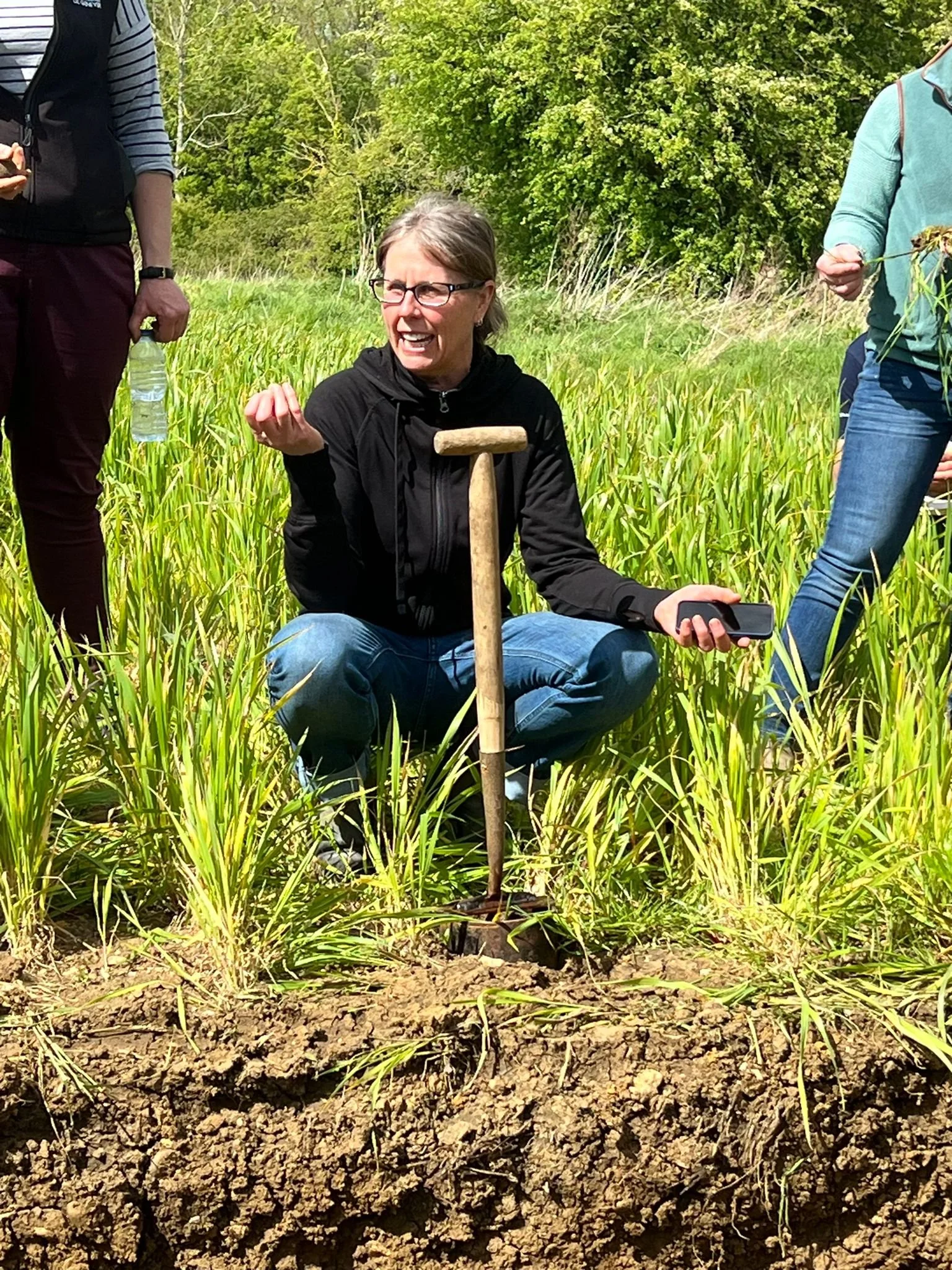 A woman crouching in a green field with tall grass, holding a smartphone, while a soil sampling instrument is positioned in the ground. She is smiling and appears to be engaged in an outdoor activity with other people around her.