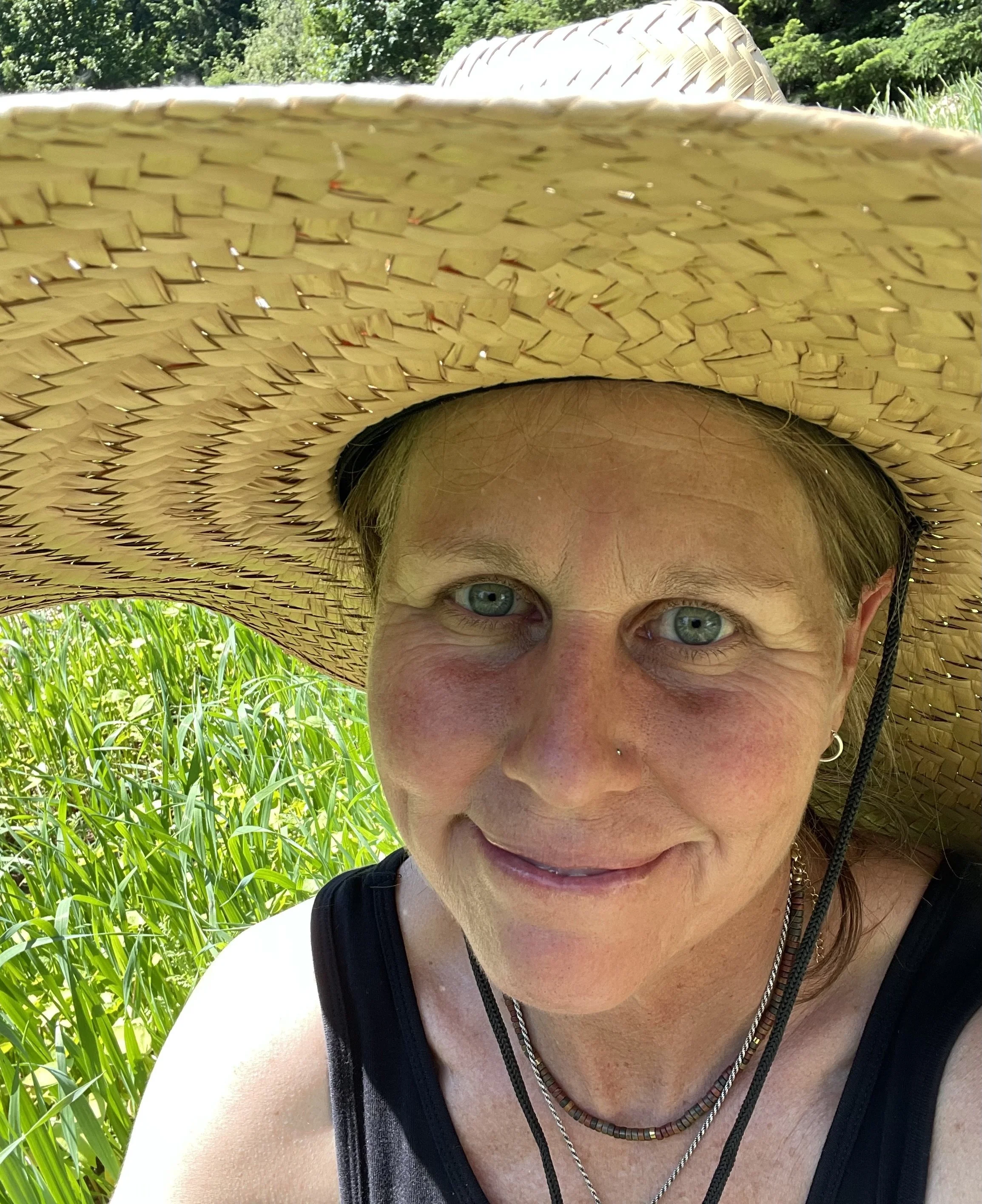 Close-up of a smiling woman wearing a wide-brimmed straw hat, standing outdoors in a field of tall green grass.