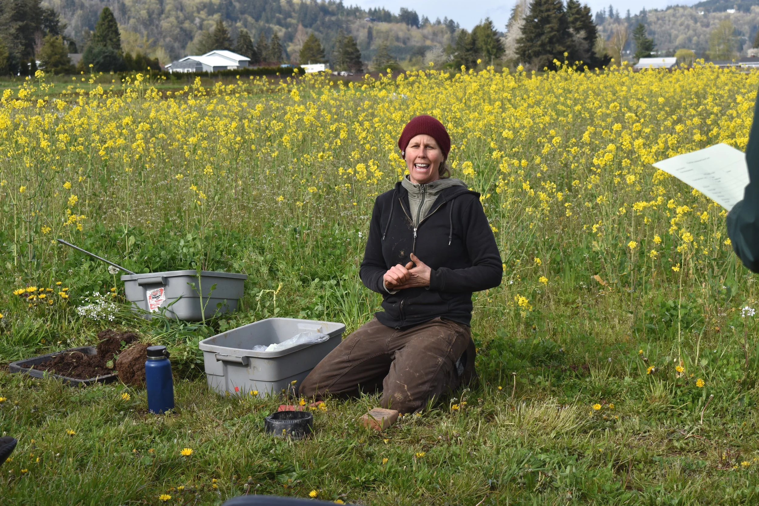A woman kneeling in a field of yellow flowers, speaking. She is wearing a red beanie, black jacket, and brown pants, with gardening supplies nearby.