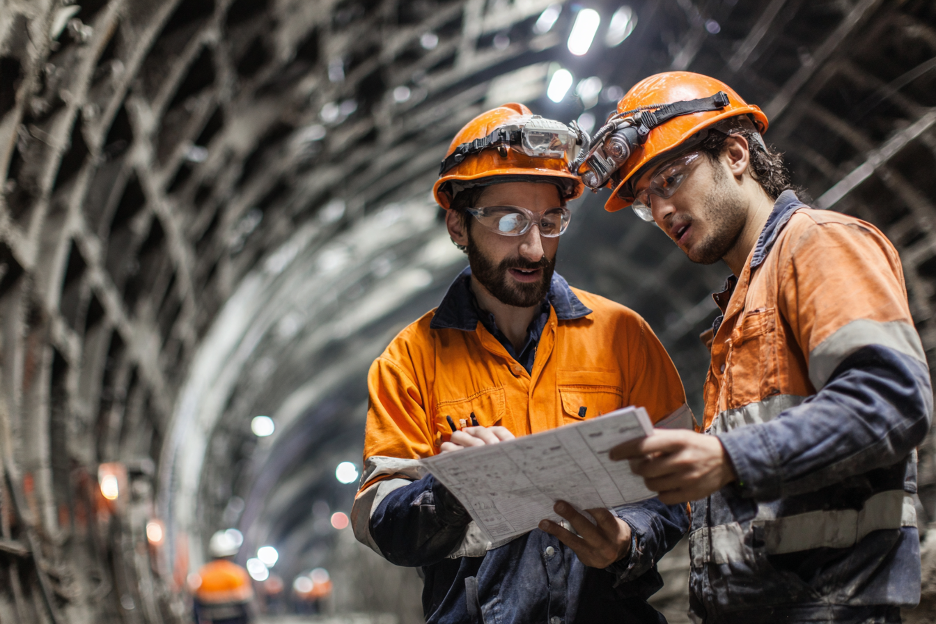 Two underground miners in orange safety gear and helmets reviewing a blueprint inside a tunnel