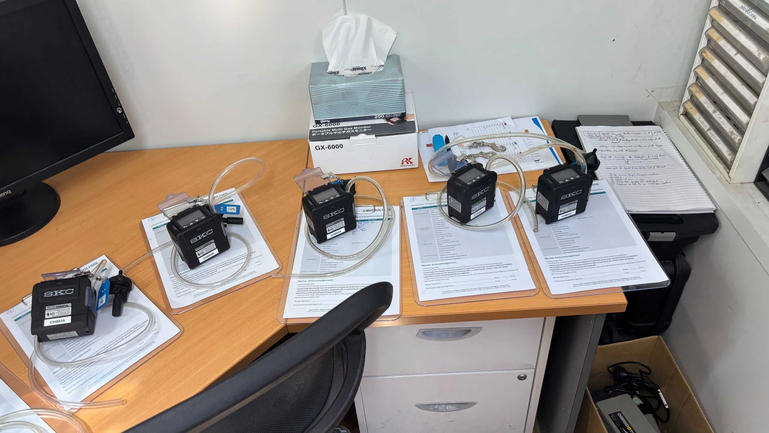 Medical equipment, including three black SKC gas monitors for Silica monitoring with attached tubing, placed on documents on a desk with a computer monitor, a tissue box, and stacks of paper and files.