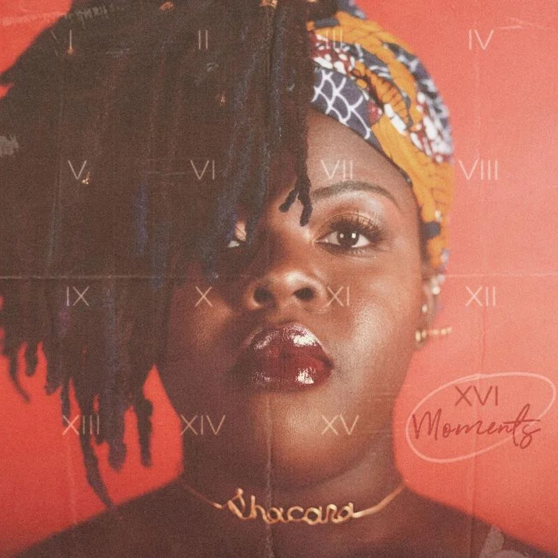 Close-up portrait of a woman with styled dreadlocks partially covering one eye, wearing a colorful headwrap, glossy lipstick, and a necklace with the word 'Chocola'. She has small earrings and a confident expression with a red background.