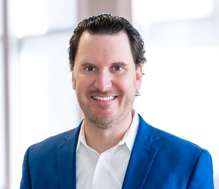 A smiling man with dark hair wearing a blue suit and white shirt, standing indoors with a blurred window background.