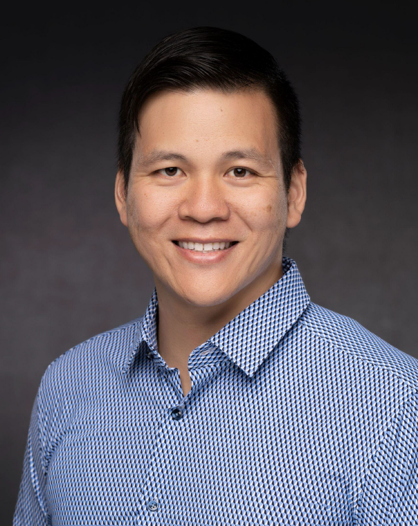 Portrait of a man with short dark hair, smiling, wearing a blue and white checkered shirt, against a dark gray background.