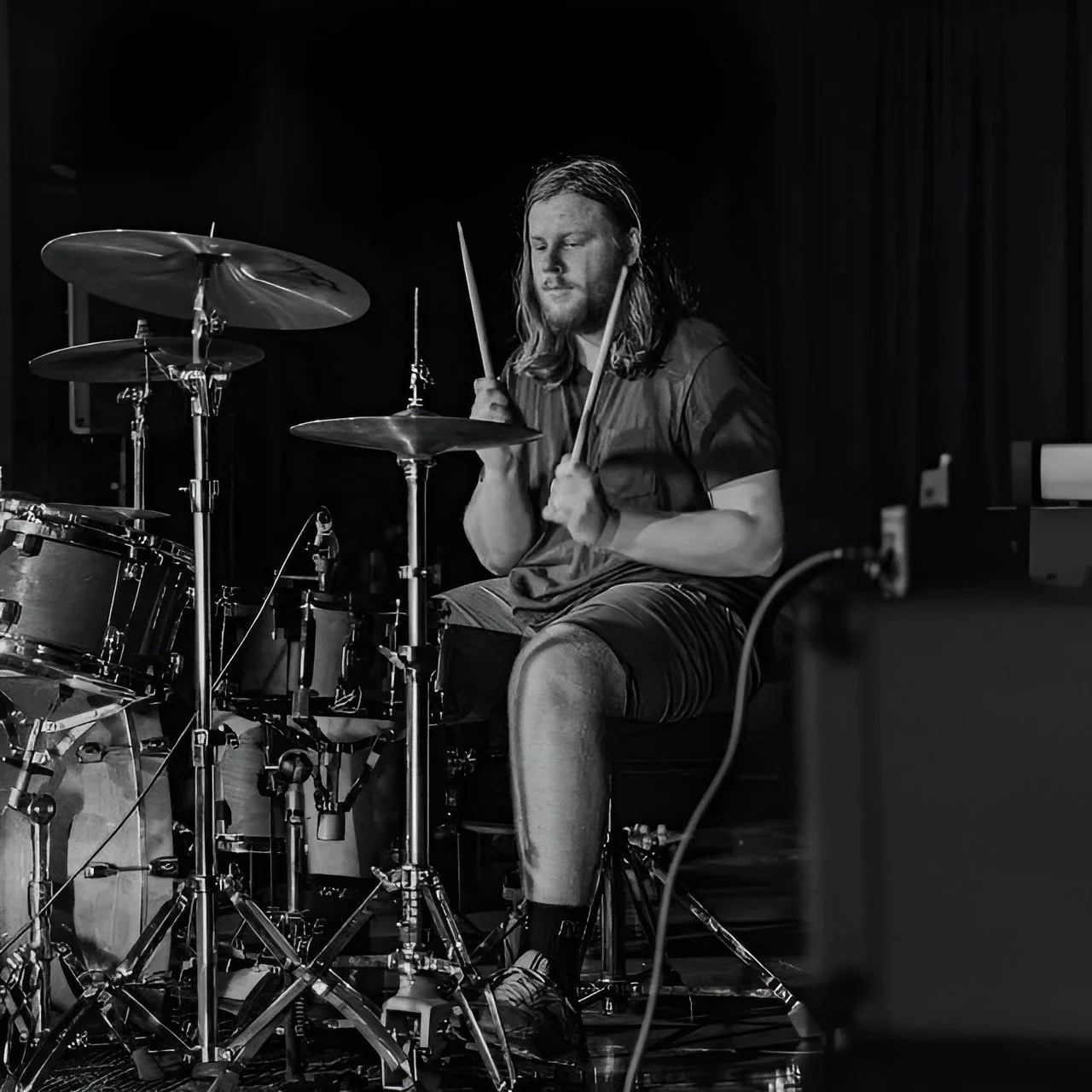 A man with long hair and a beard playing the drums on a stage, captured in black and white.