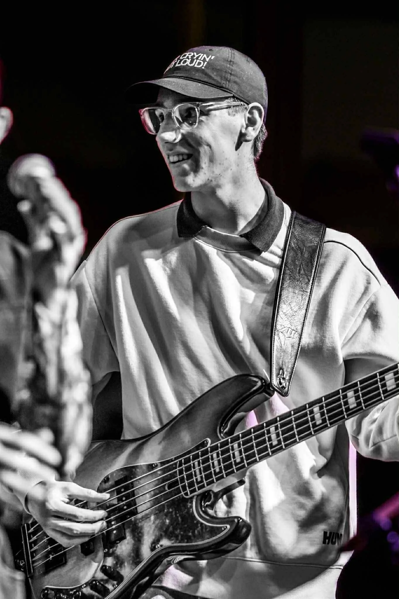 A young man smiling while wearing glasses and a cap with the text 'Cryin' Out Loud!' playing an electric bass guitar in a dark environment.