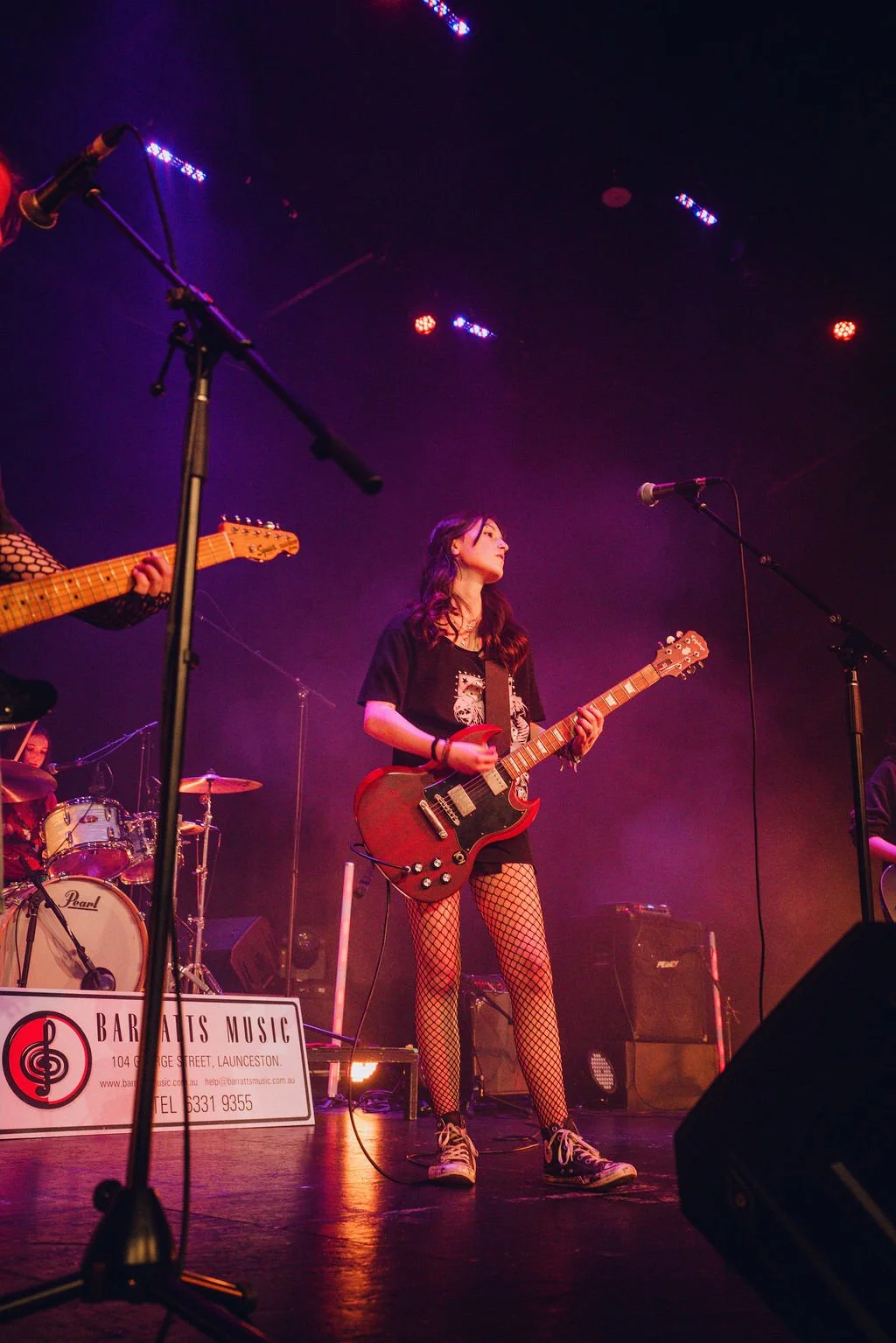 A young woman playing an electric guitar on stage during a live music performance, with purple and orange stage lights and a drum set in the background.