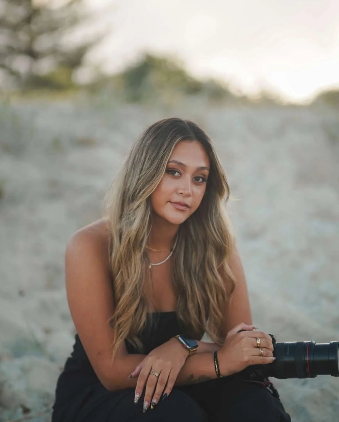A woman with long wavy hair sits on the sand at the beach, holding a camera, wearing a black top, layered necklaces, a smartwatch, and rings.