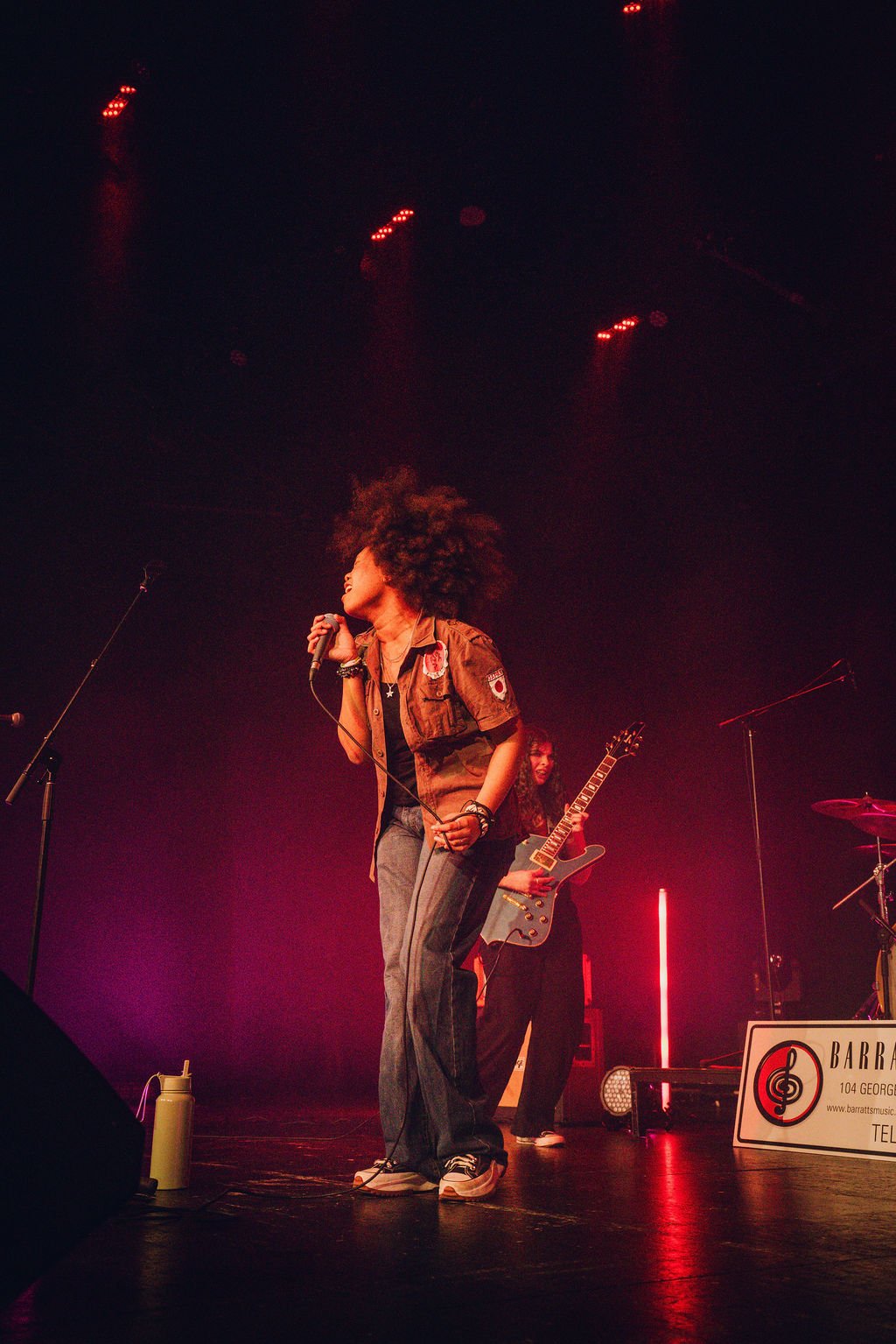 A female singer with curly hair performs on stage with a microphone, accompanied by a guitarist in the background. Stage lighting creates a dark, moody atmosphere with red and purple accents.