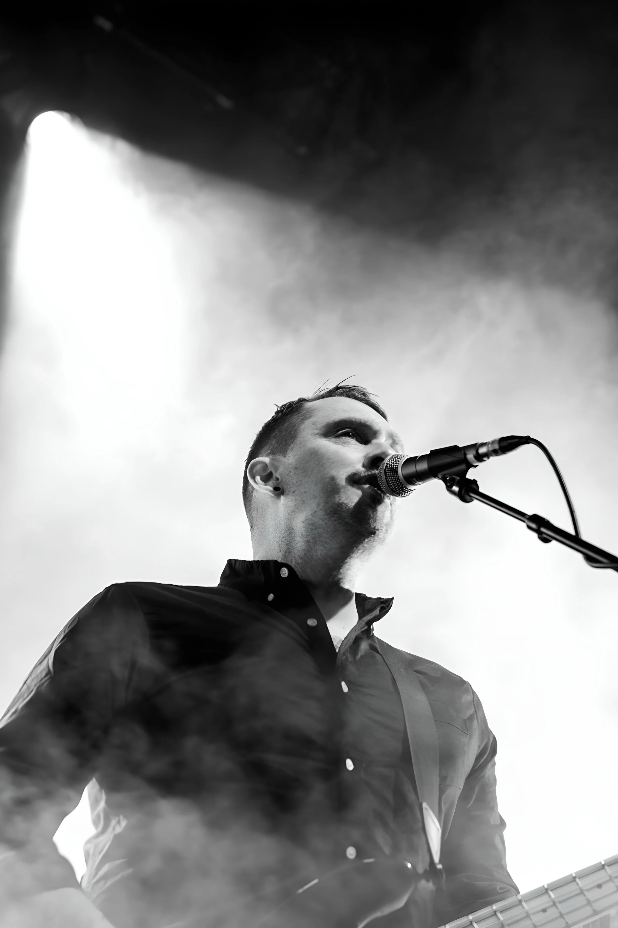 A male musician singing into a microphone while playing a guitar, with stage lighting and fog effects in black and white.