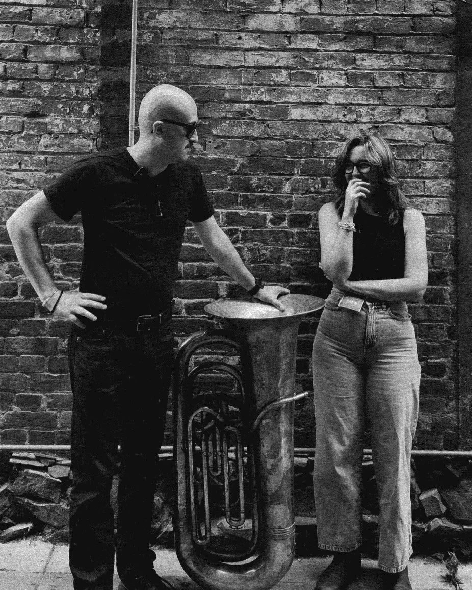 A man and woman standing against a brick wall with a tuba placed between them. The man has a shaved head, glasses, and is wearing a black shirt, while the woman has wavy hair, glasses, and is wearing a sleeveless top and jeans, both appear to be in a casual conversation.