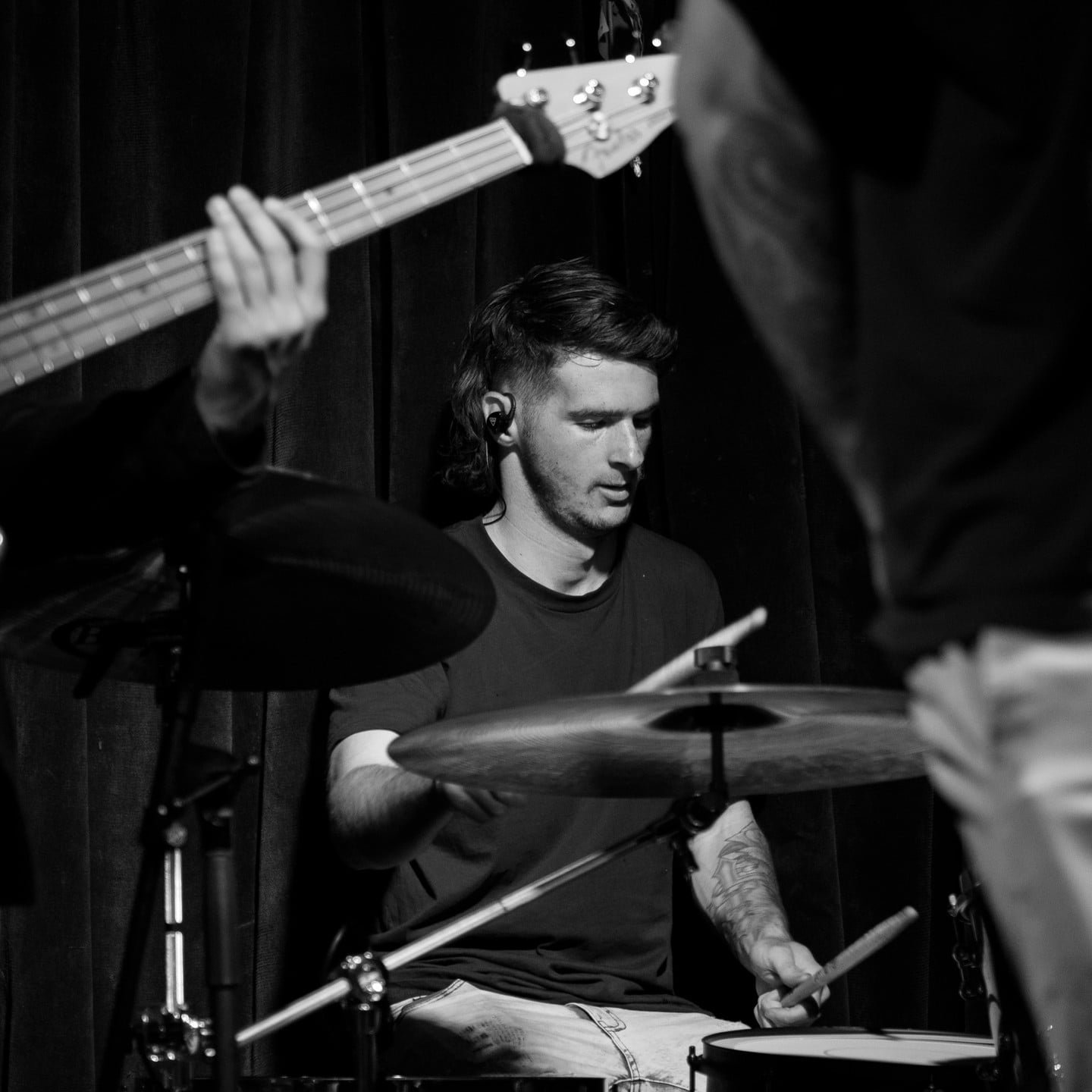 Black and white photo of a young man playing drums on a stage, with another musician playing the guitar partially visible in the foreground.