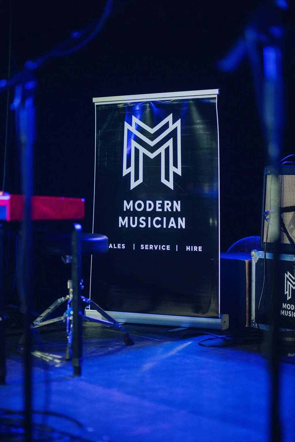 Stage banner with a modern logo and text reading 'Modern Musician' and features 'Sales, Service, Hire' underneath, with music equipment nearby.