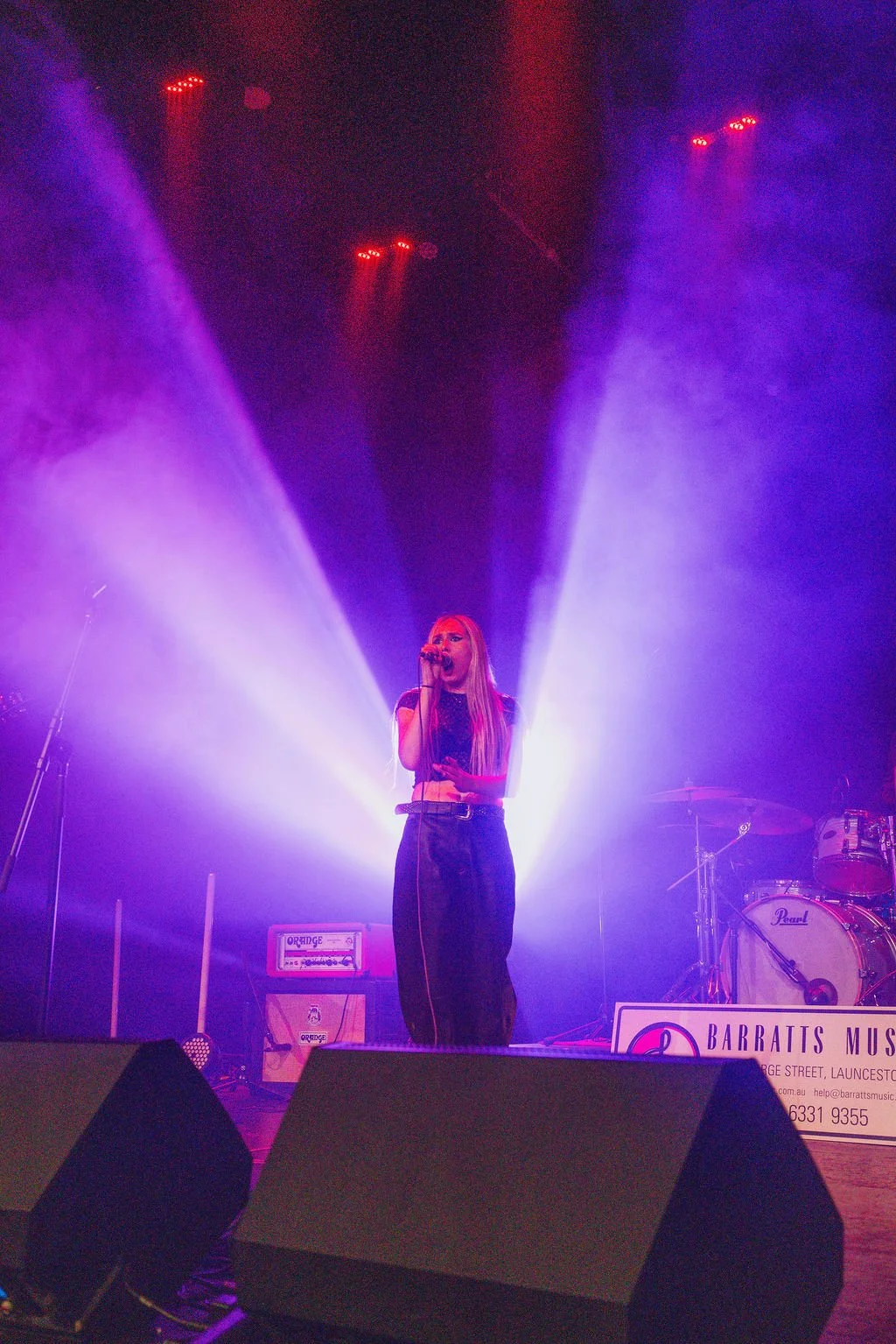 A female singer performing on stage with purple and blue lighting and smoke effects, holding a microphone. There is musical equipment and a sign that reads 'BARRAITS MUSIC' in the background.