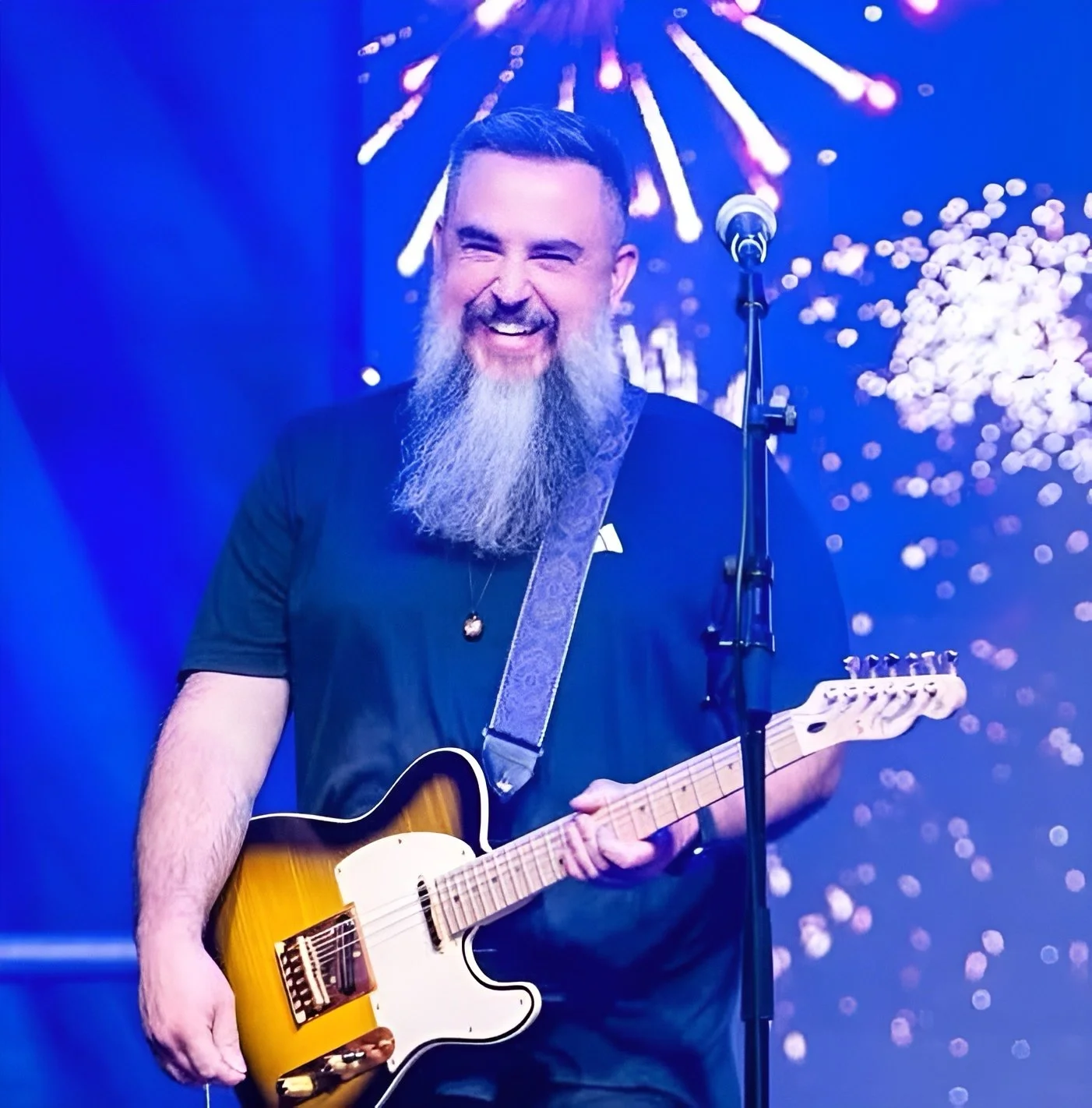 A man with a beard playing an electric guitar on stage with fireworks display in the background.