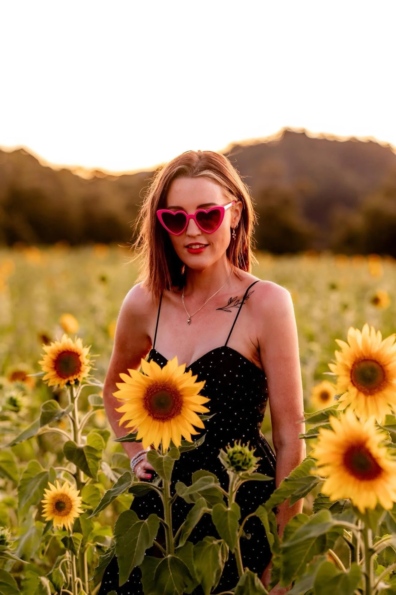 A woman with red hair, pink heart-shaped sunglasses, and a black dress with white polka dots standing in a sunflower field during sunset.