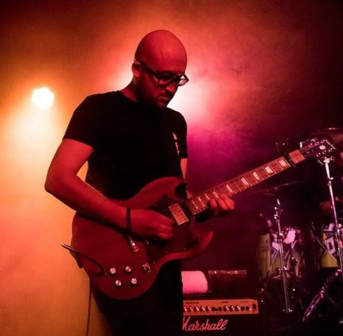 A musician with glasses playing an electric guitar on stage with colored lighting.