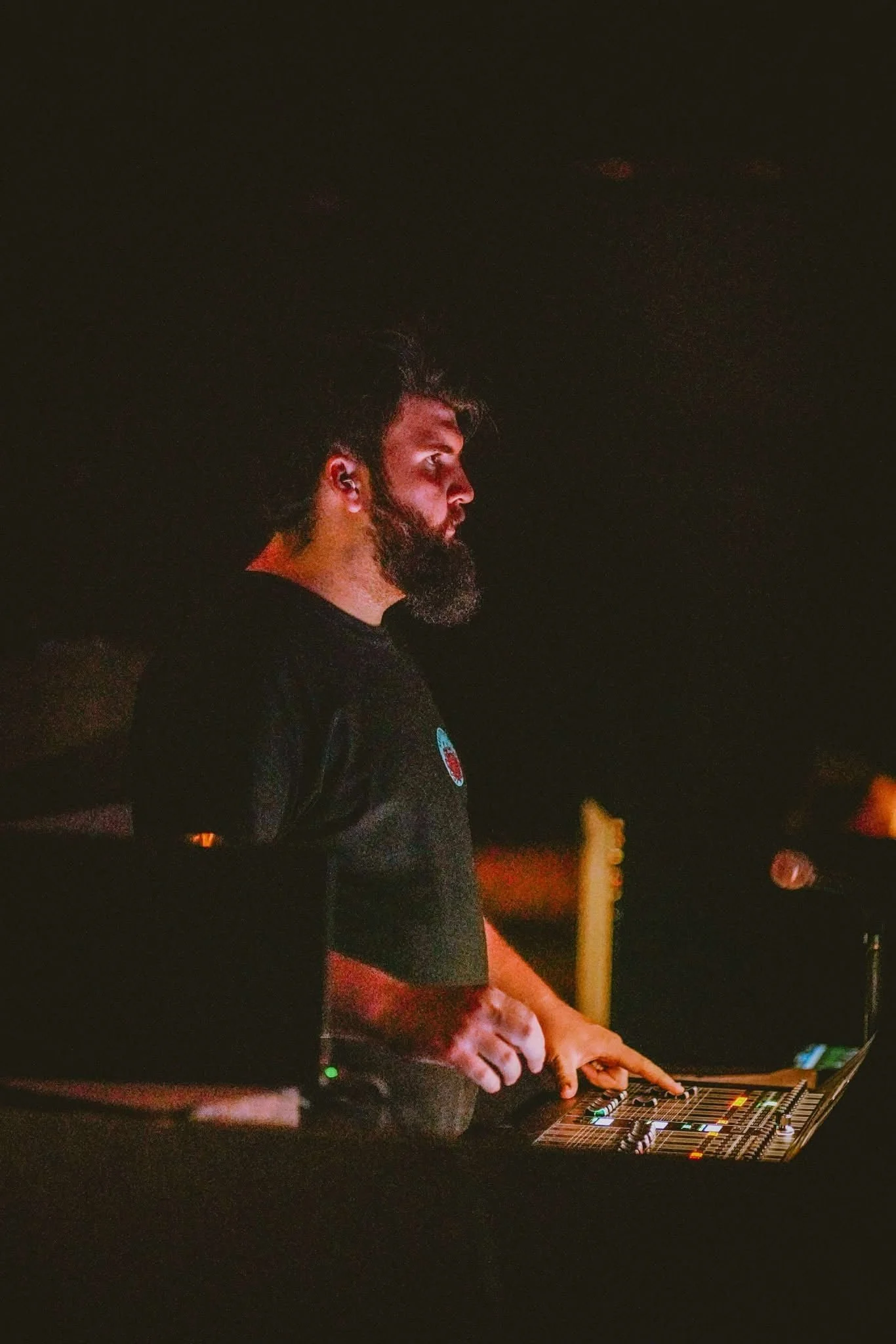 A bearded man with curly hair operating an audio mixing console in a dark setting, possibly at a music event or studio.