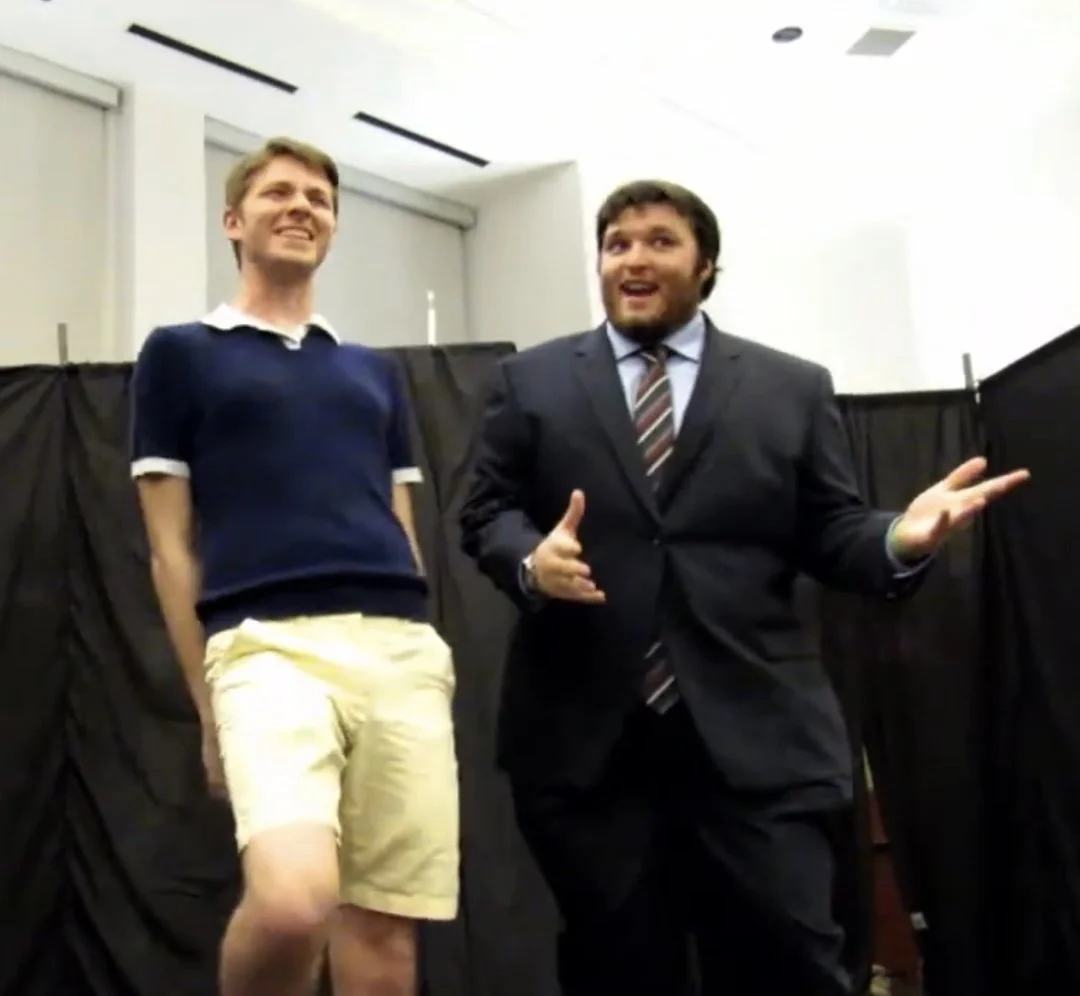 Two men are standing and talking to each other in a room with black curtains, one wearing a casual navy shirt and shorts, the other in a business suit with a striped tie.