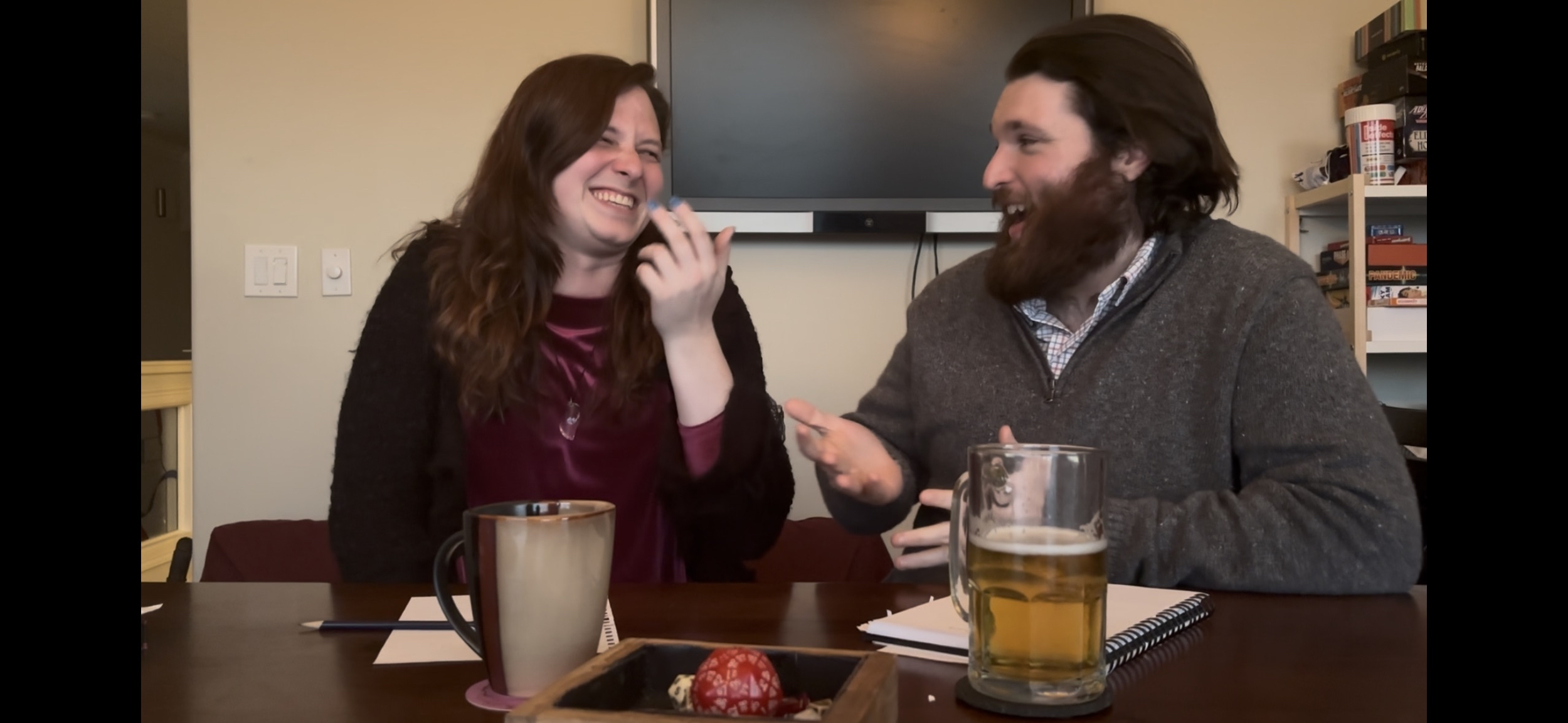 A woman and a man laughing and talking at a table with drinks, notebooks, and a decorative egg in a cozy room.