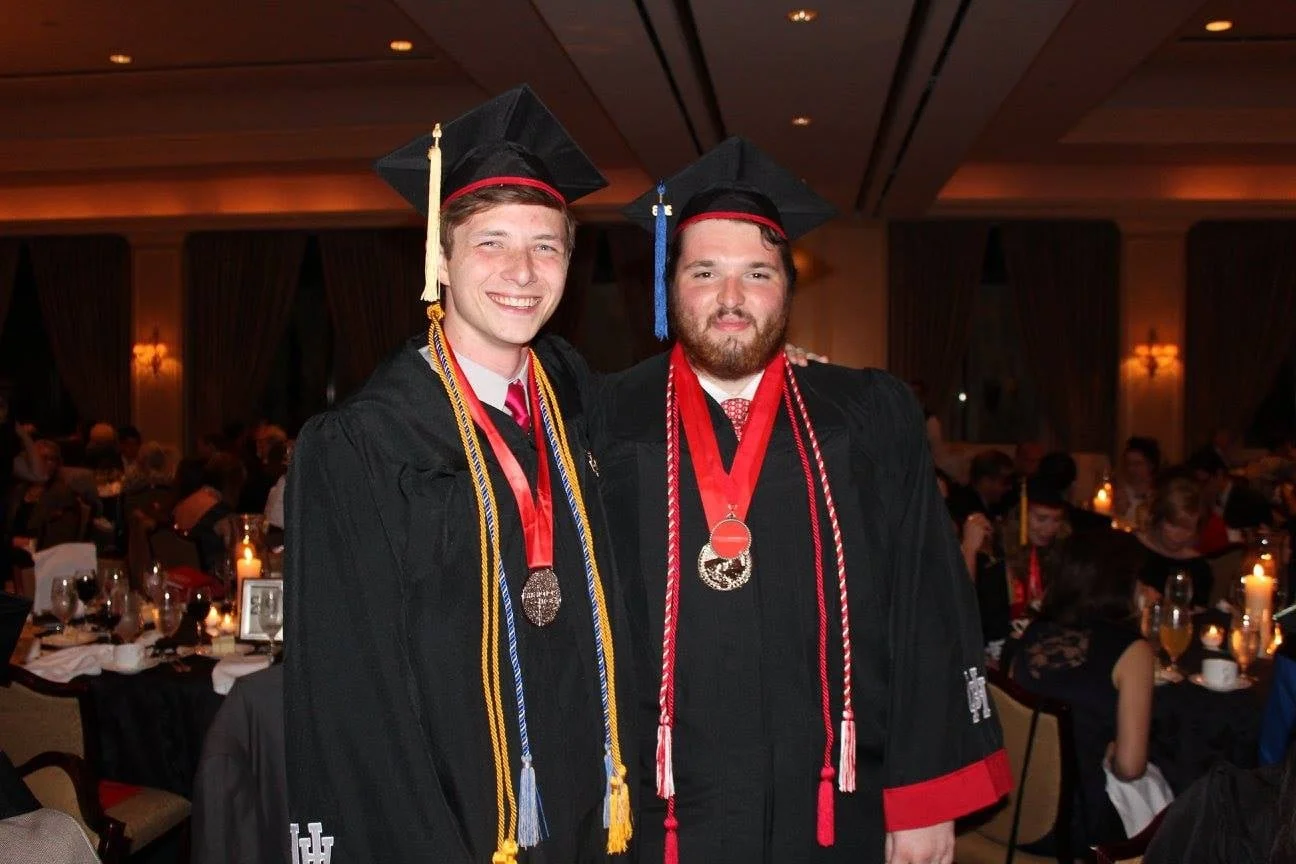 Two young men in black graduation gowns and caps with tassels, wearing medals around their necks, smiling and standing close together at a graduation ceremony in a decorated banquet hall.