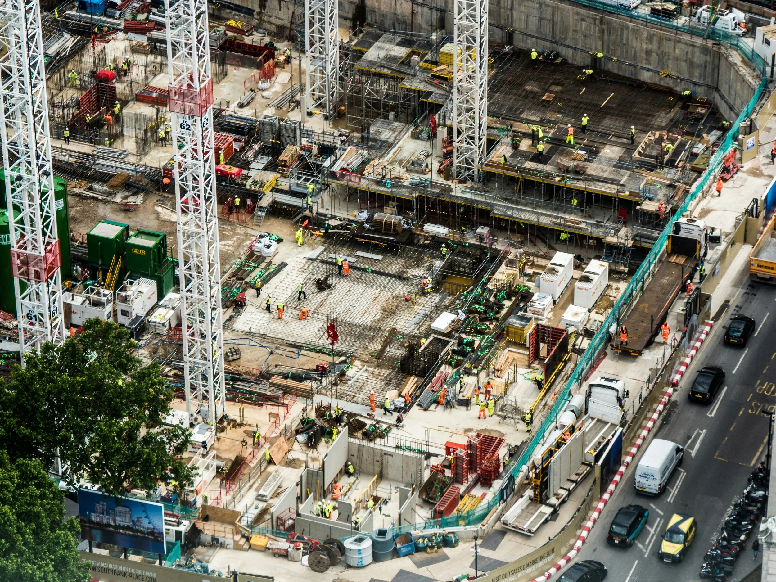 Aerial view of a busy construction site with workers, cranes, and building materials, alongside a road with vehicles.