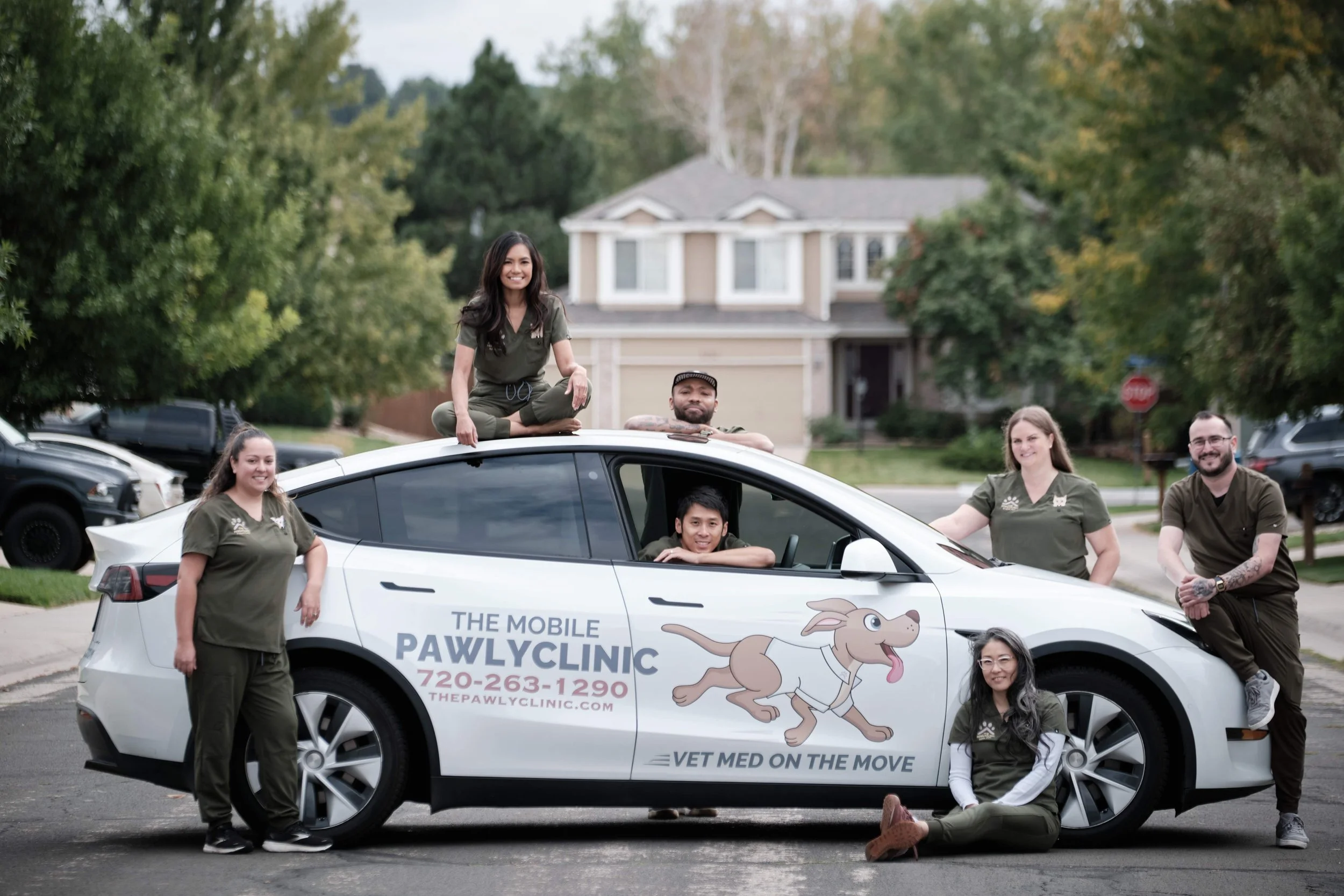 Group of six people posing around a white Tesla car with 'PAWLYCLINIC' and a cartoon dog graphic, front photo in a suburban neighborhood, some inside the car, some standing or sitting outside.