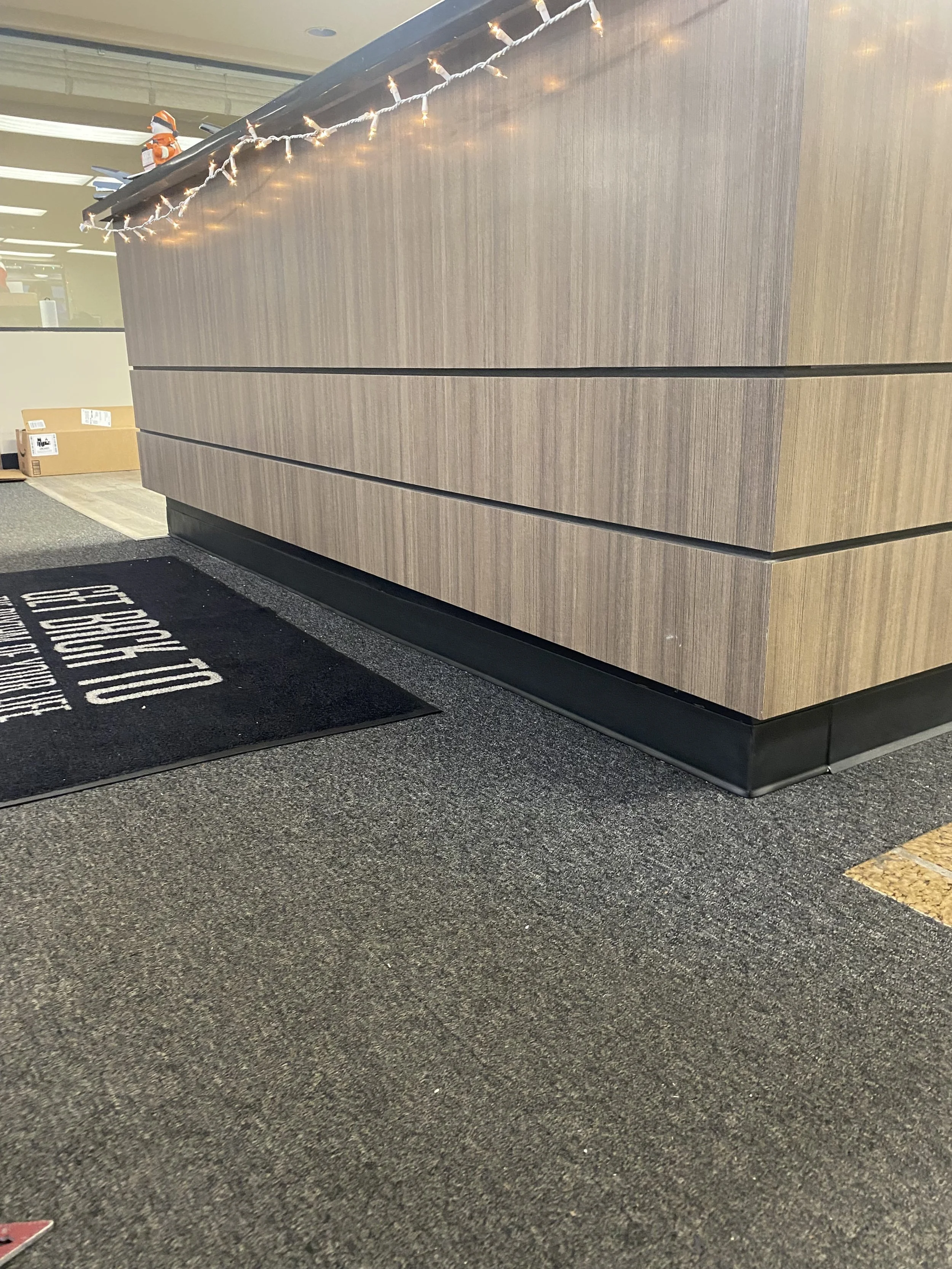 Close-up of a reception desk with string lights and a decorative item on top. There is a black mat on the gray carpet in front of the desk, with white text that says 'EMPLOYEE OF THE MONTH'.