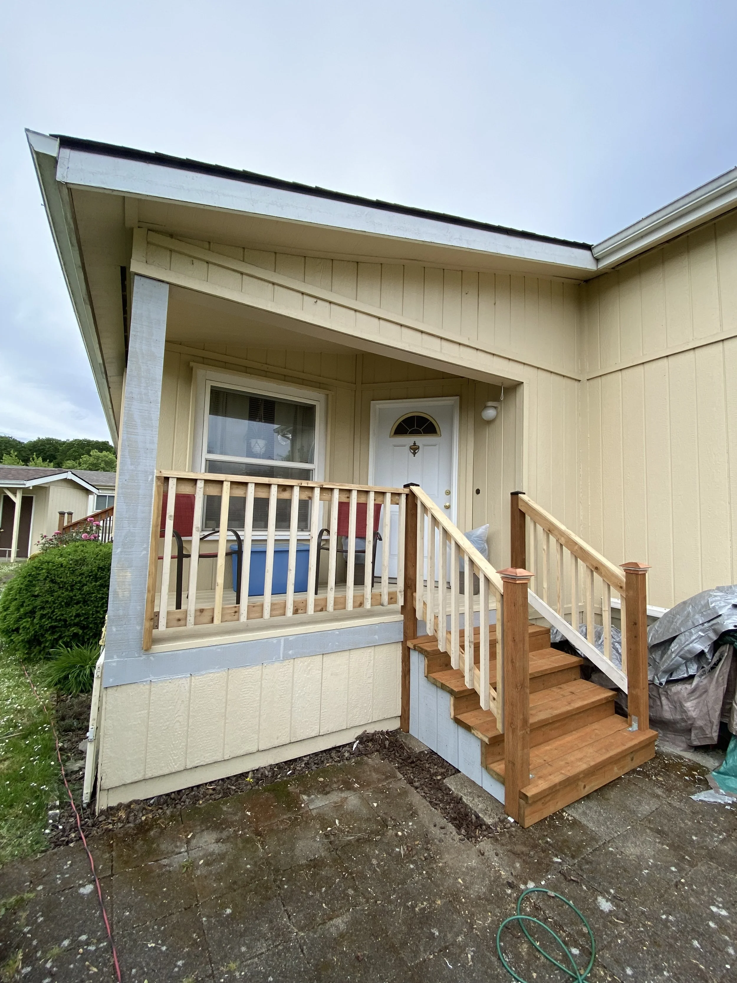 Front porch under construction with new wooden stairs and railing, attached to a yellow house with a white door and window.