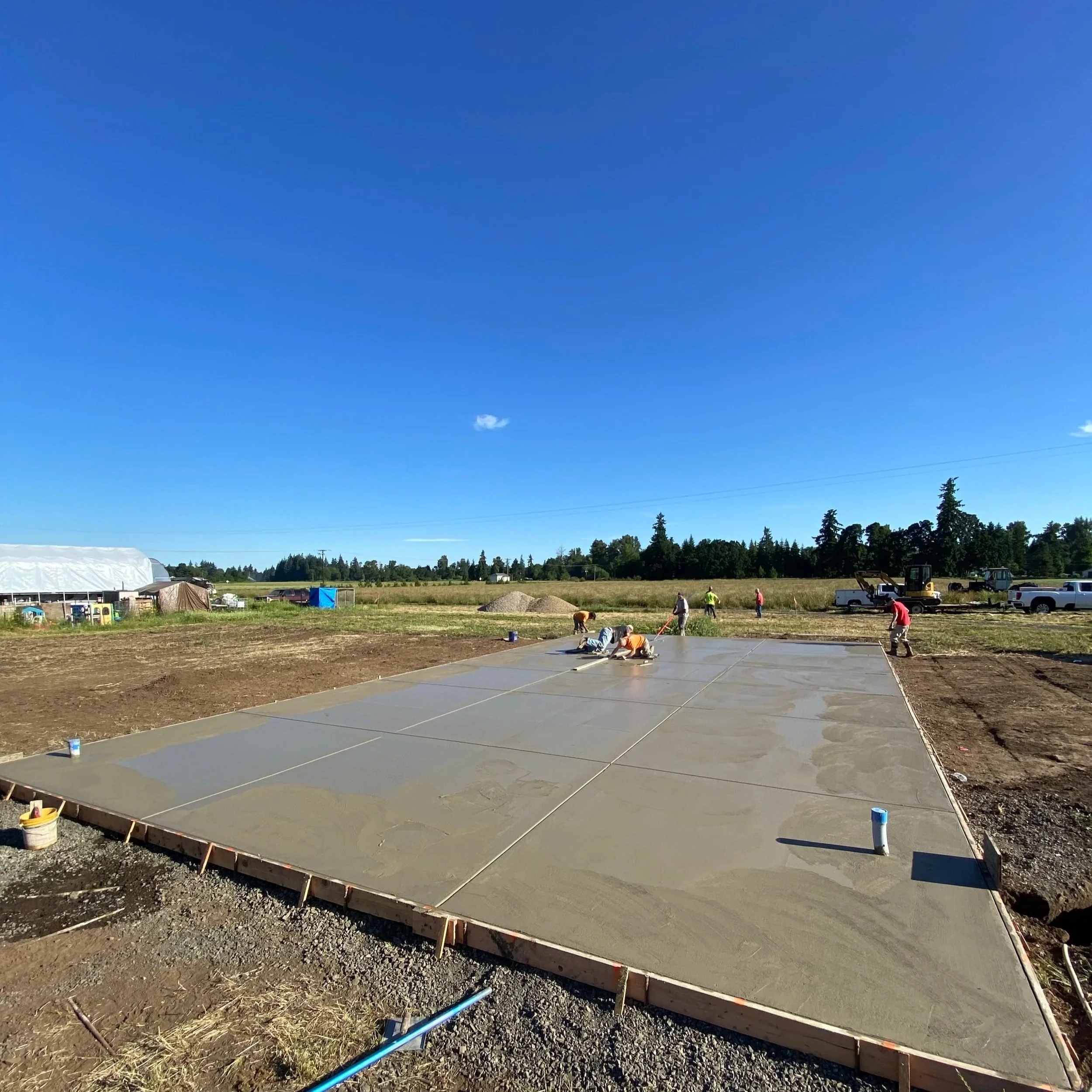 Workers pouring and smoothing concrete on a construction site with open land and trees in the background under a clear blue sky.