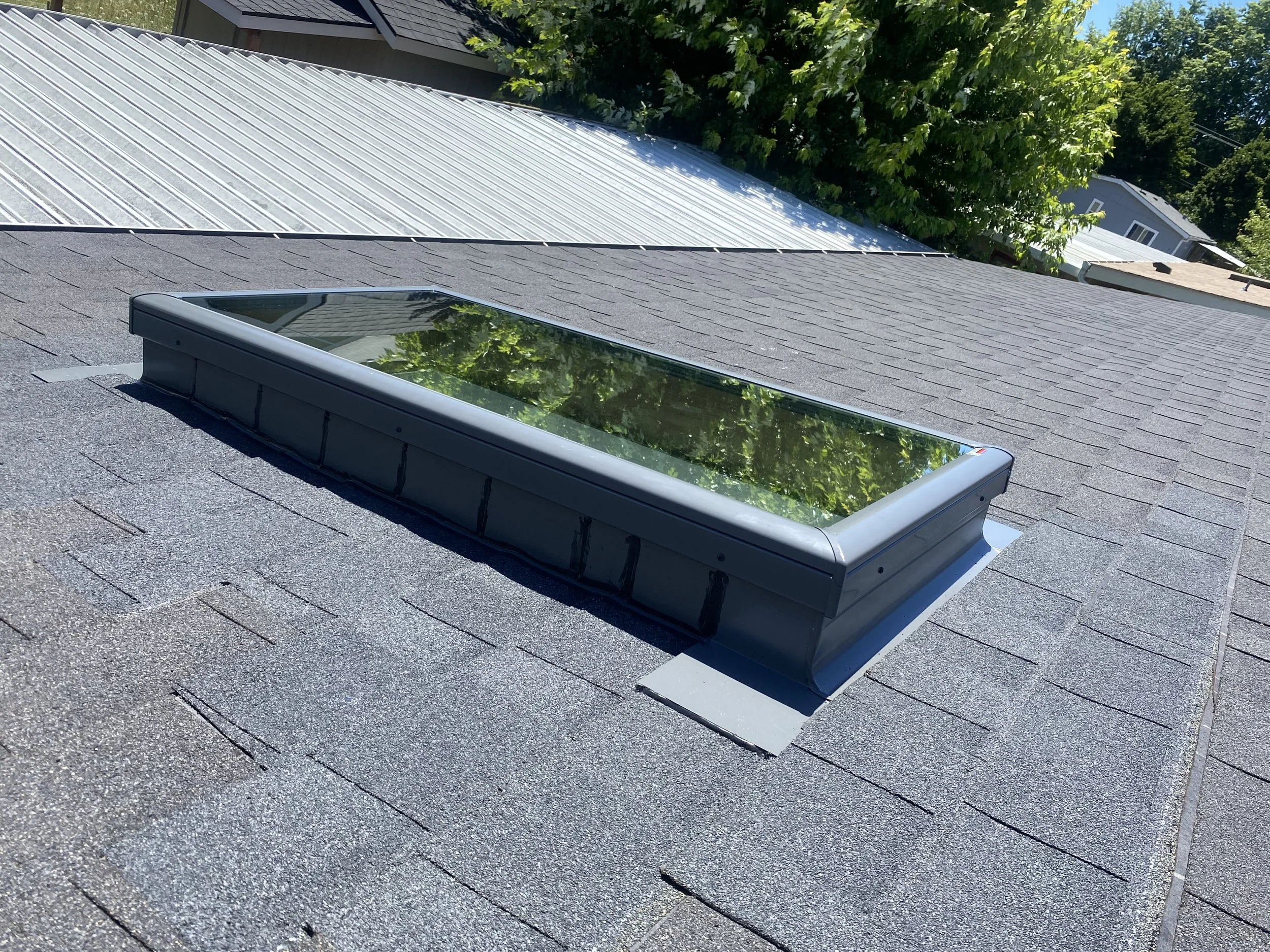 A rectangular skylight with a glass window installed on a shingled roof, with green trees and neighboring rooftops visible in the background.