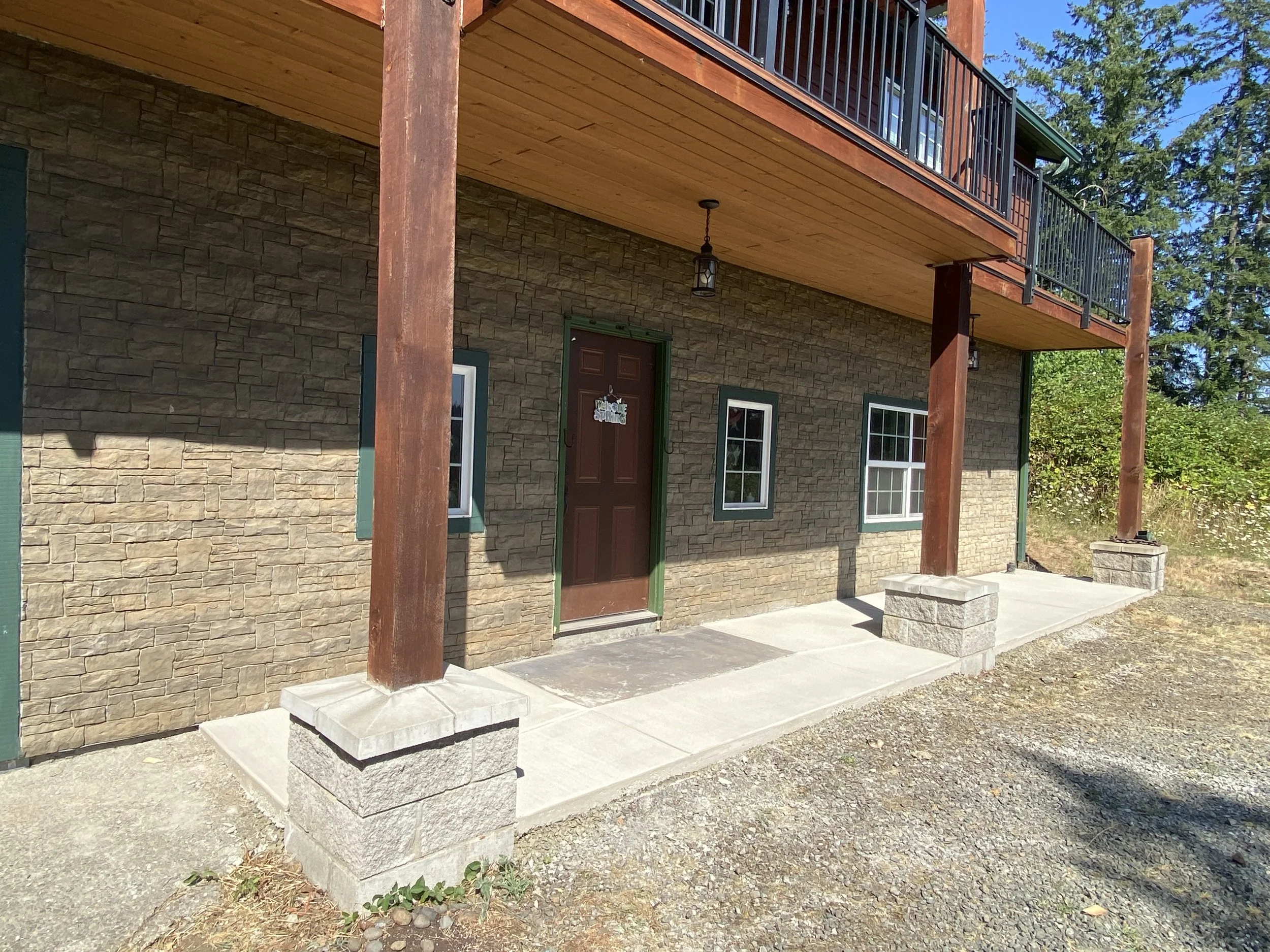 Front view of a house with a stone facade, green window frames, a brown door, concrete porch steps, and a balcony with black railing supported by wooden posts.