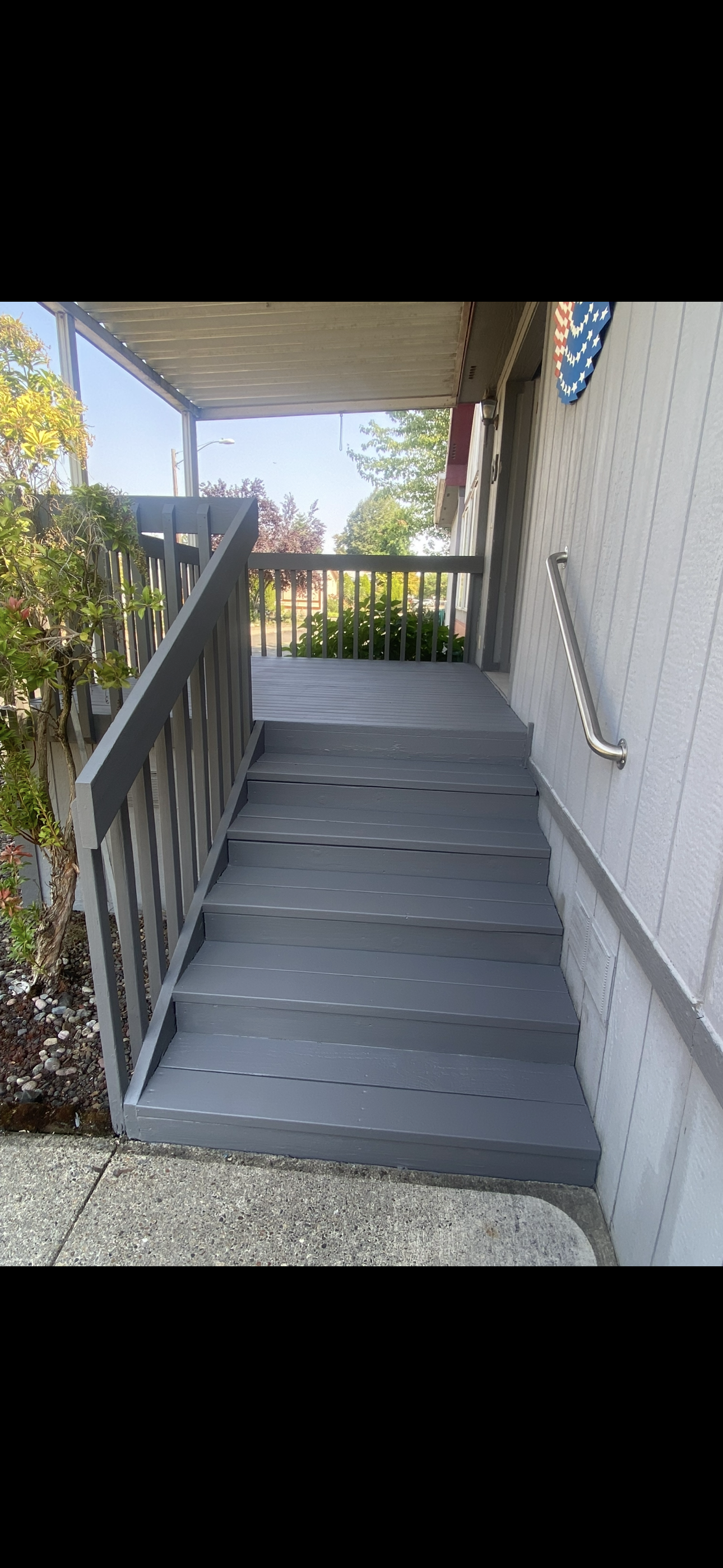 A backyard with a newly built wooden deck attached to a yellow house with white trim. The deck has a small staircase and railing, and there are potted plants and outdoor furniture nearby under a clear blue sky.