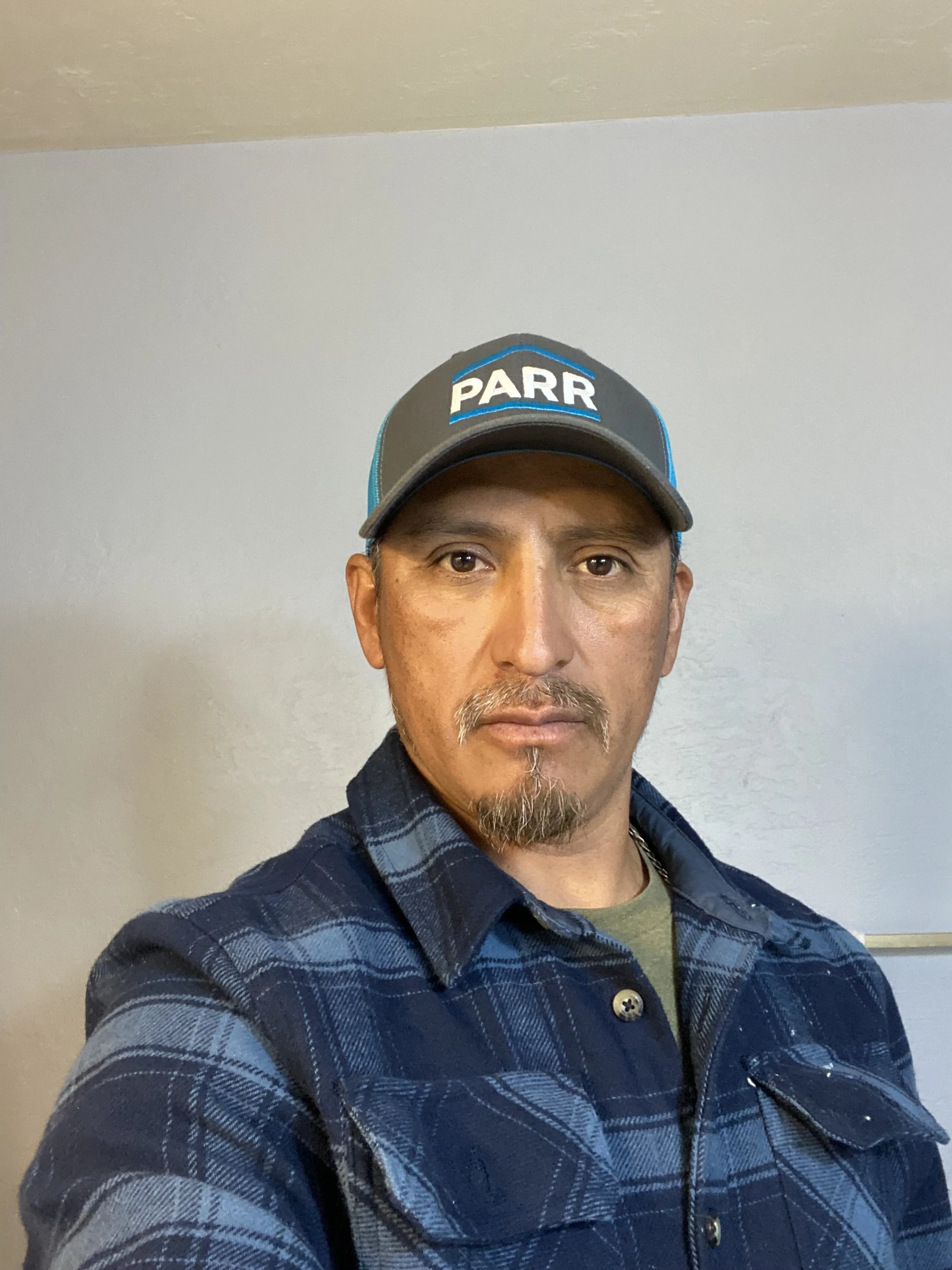A man wearing a blue and black plaid shirt and a black cap with the word 'PARR' written on it, standing against a plain light-colored wall.