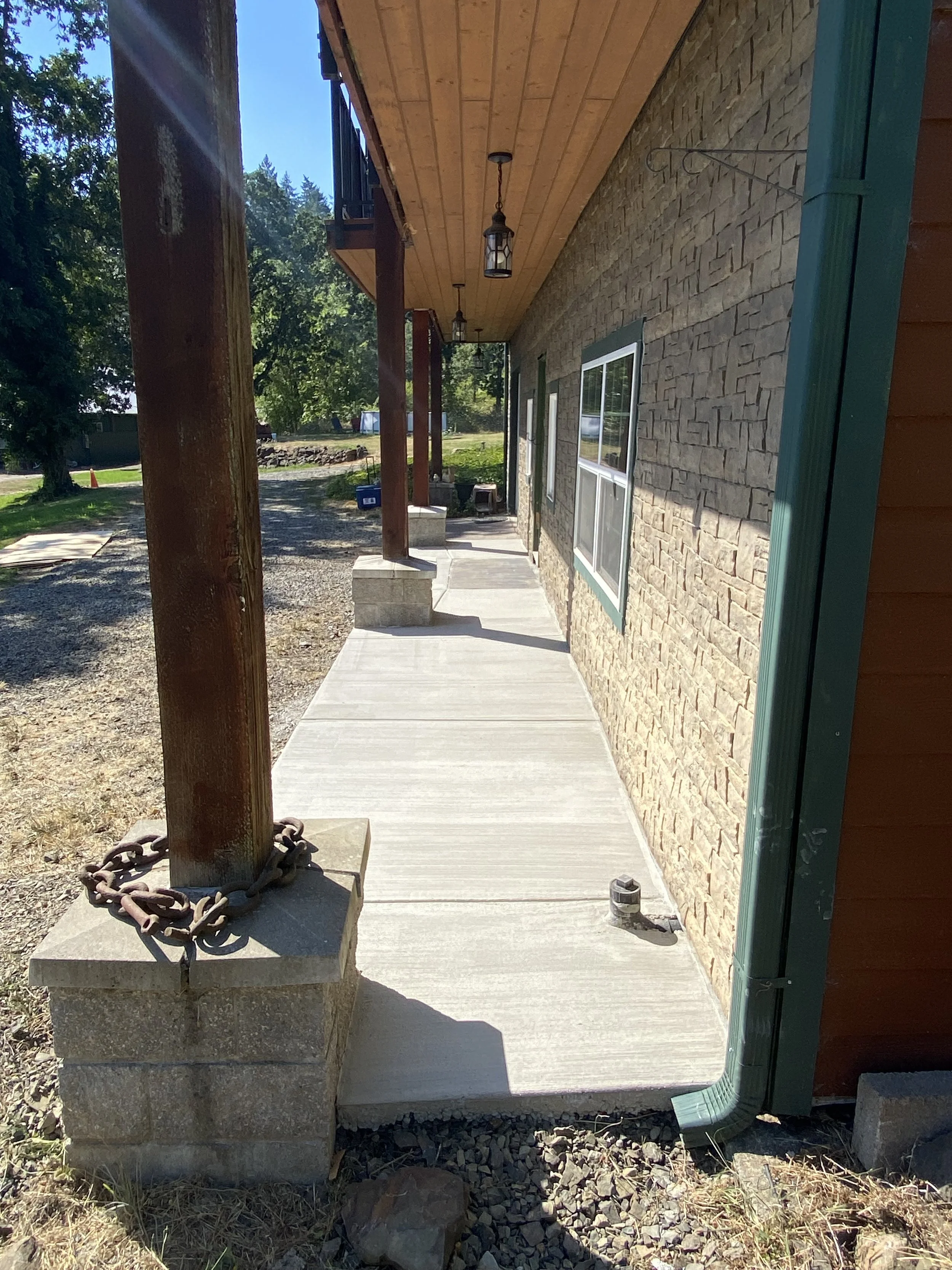 View of a concrete porch with support posts, attached to a building with brick exterior wall and windows. Hanging light fixtures and a green rain gutter are visible along the porch.