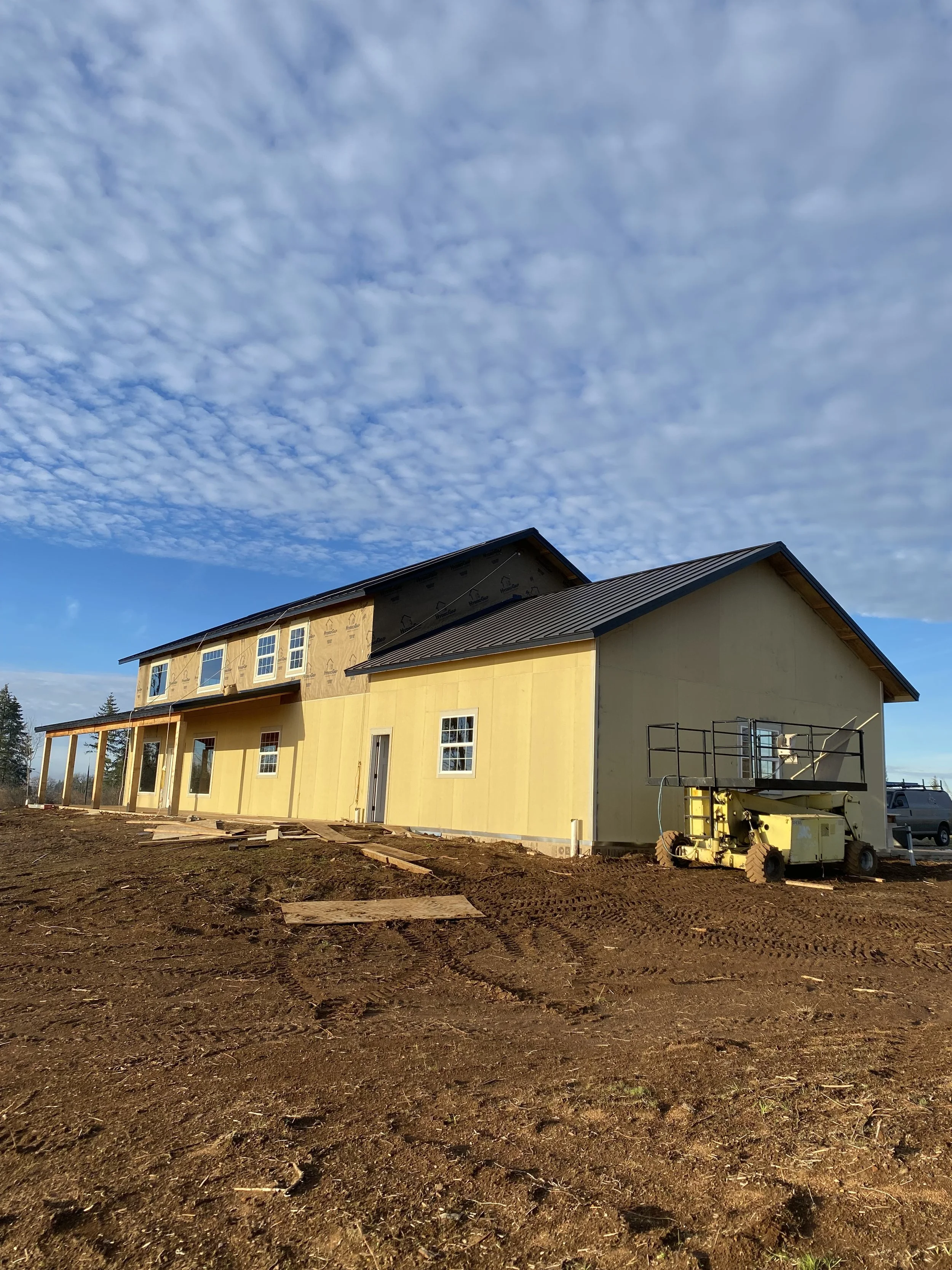 Under-construction house with yellow siding and black roof, construction equipment, and dirt ground, under a cloudy sky.