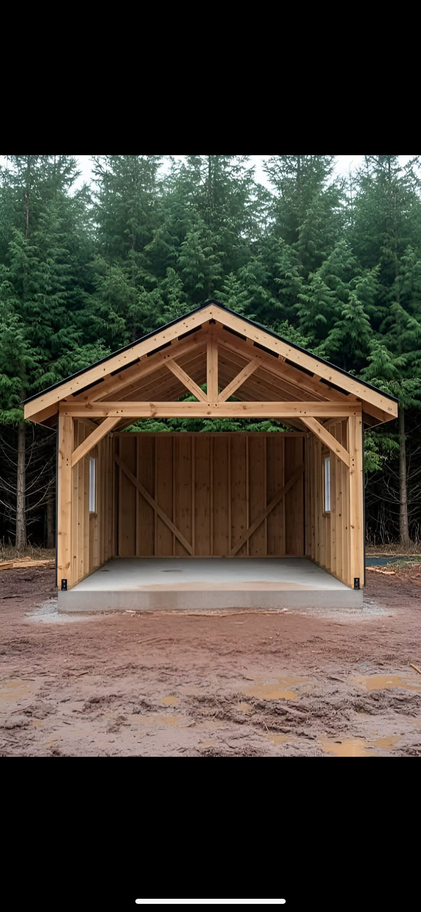 Newly constructed wooden garage or shed with a concrete slab foundation, situated outdoors with a forest of evergreen trees in the background.