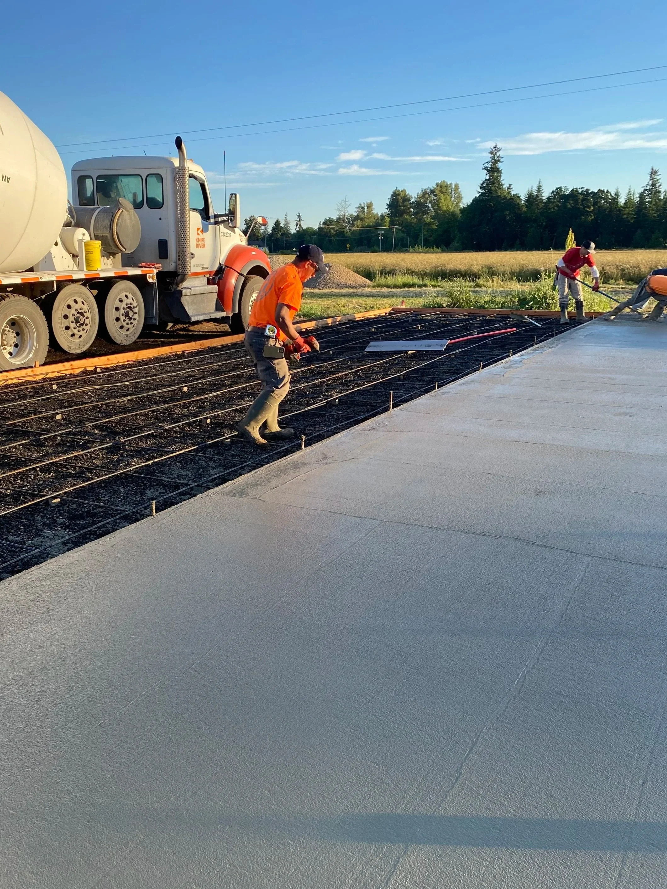 Construction workers pouring and smoothing concrete on a building's foundation with a truck parked nearby on a sunny day