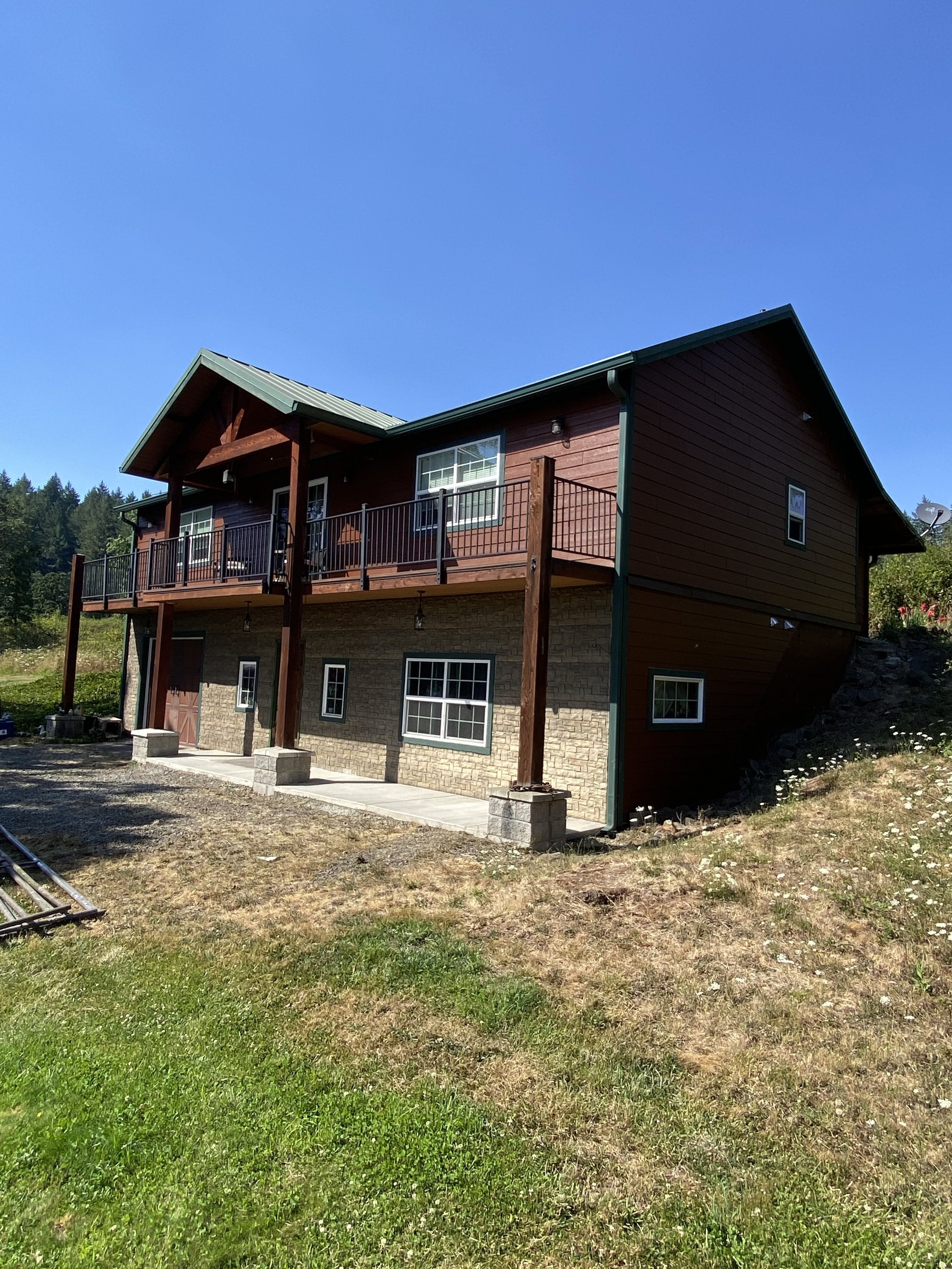 A two-story house with a wooden upper level, stone lower level, and a green metal roof, situated on a sloped grassy yard under a clear blue sky.