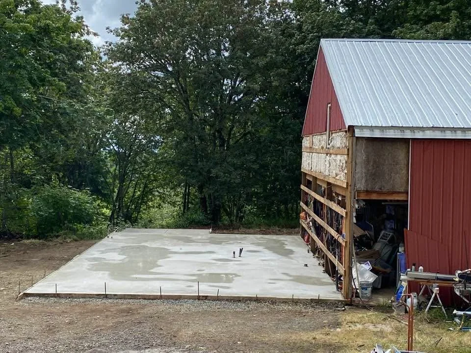 A partially built barn with a concrete slab foundation and a red metal roof, surrounded by trees.