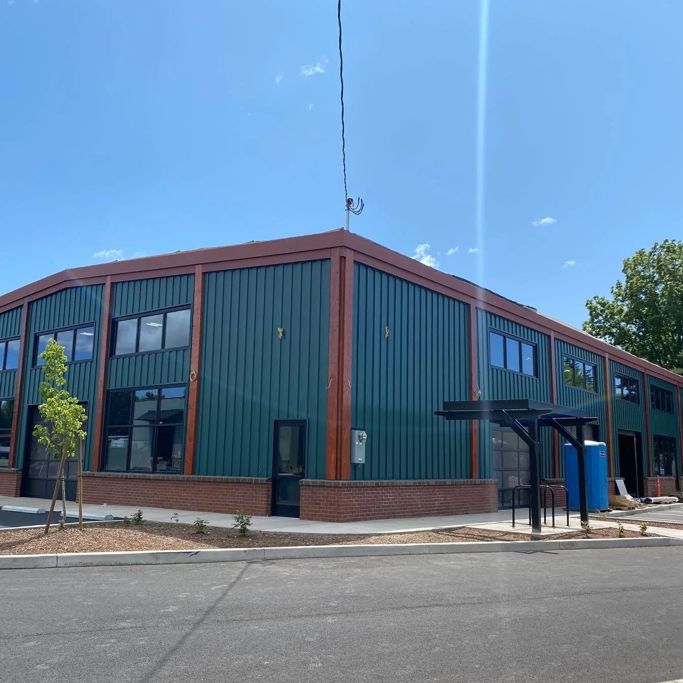 New two-story commercial building with green metal siding and brick base, featuring large windows, a small side door, and a covered bicycle rack outside under a clear blue sky.