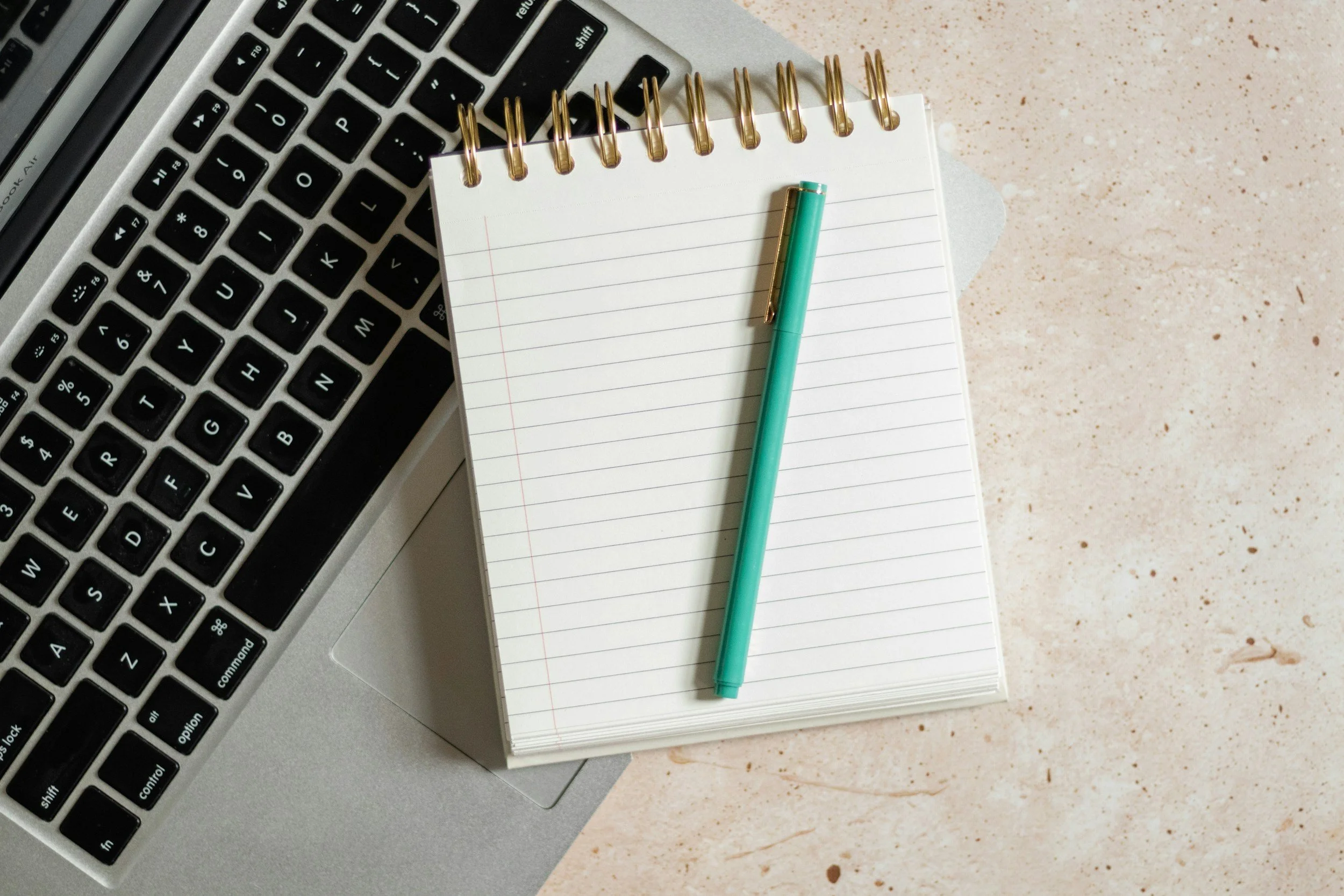A workspace featuring a silver laptop keyboard, a spiral-bound notepad with cream-colored pages, a teal pen, and a light-colored speckled surface surface.