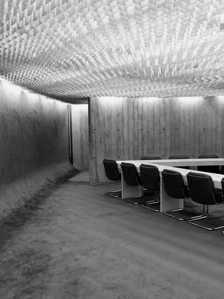 A modern conference room with wooden walls and a unique textured ceiling, featuring a long table and black chairs.