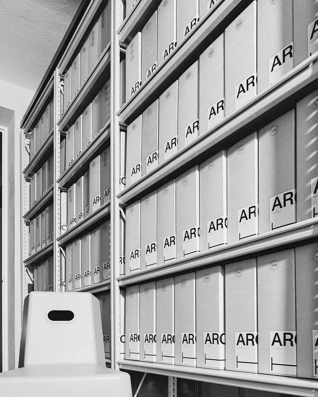 Black and white photo of metal shelves in a storage room filled with uniform boxes labeled with 'AR' and various numbers, and a white chair in front of the shelves.